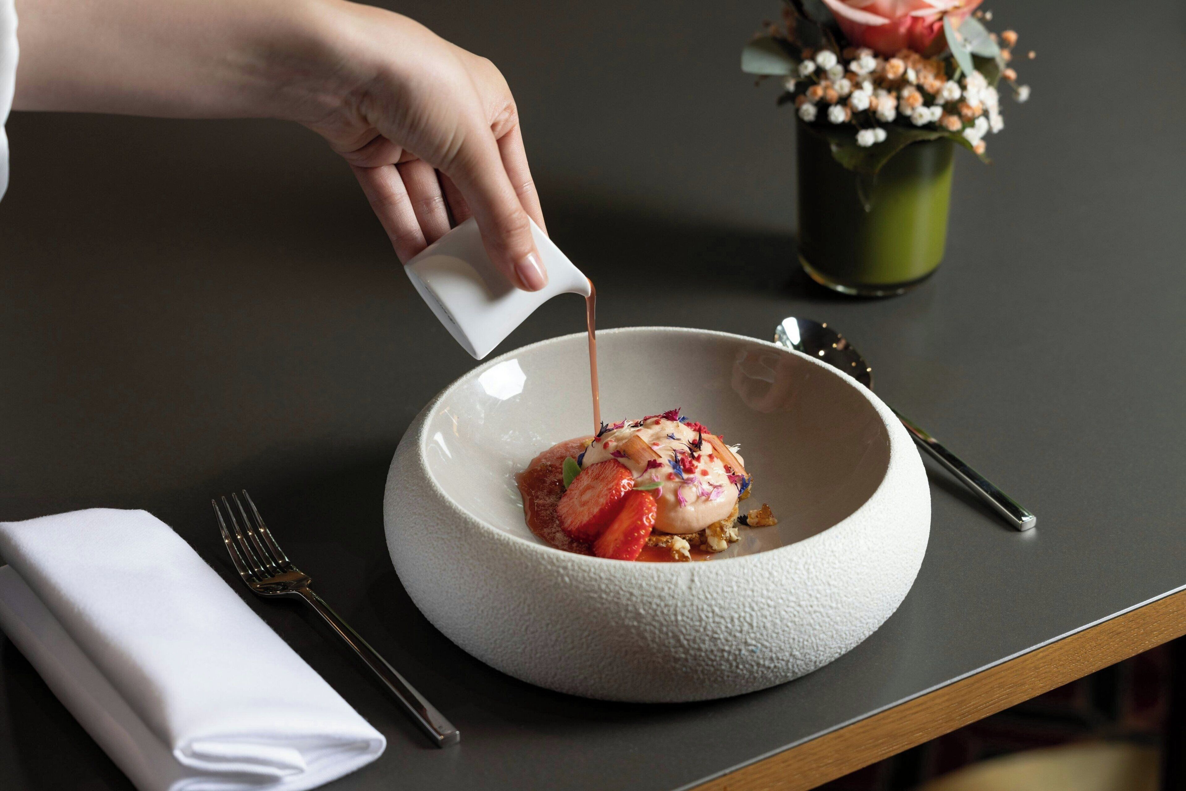 A hand pouring a liquid into a white bowl with a dessert on top.