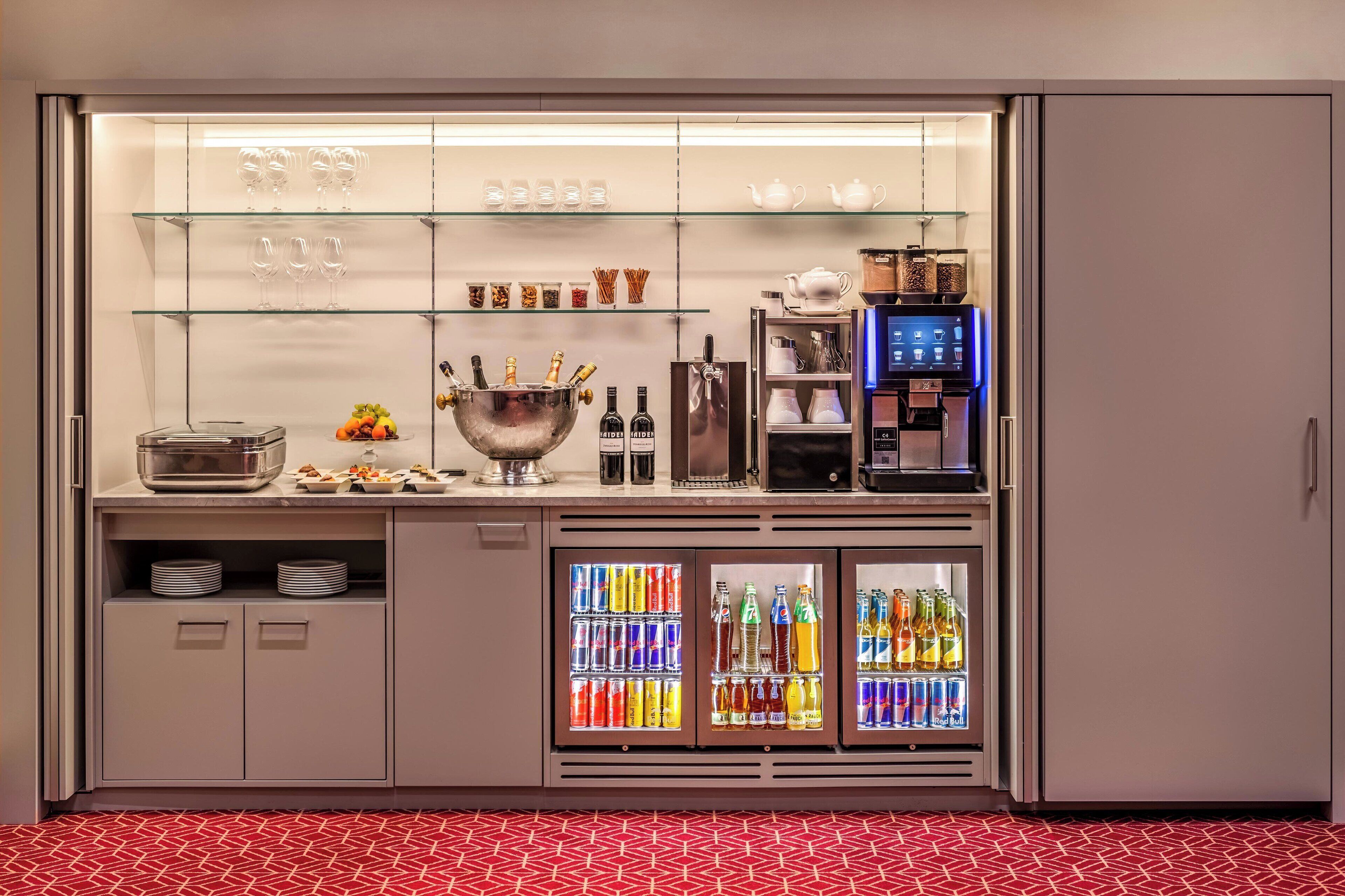 A bar area with glass shelves displaying glasses, cups, and a tea pot. Below, there are refrigerated drink dispensers and a bowl with fruits and snacks.