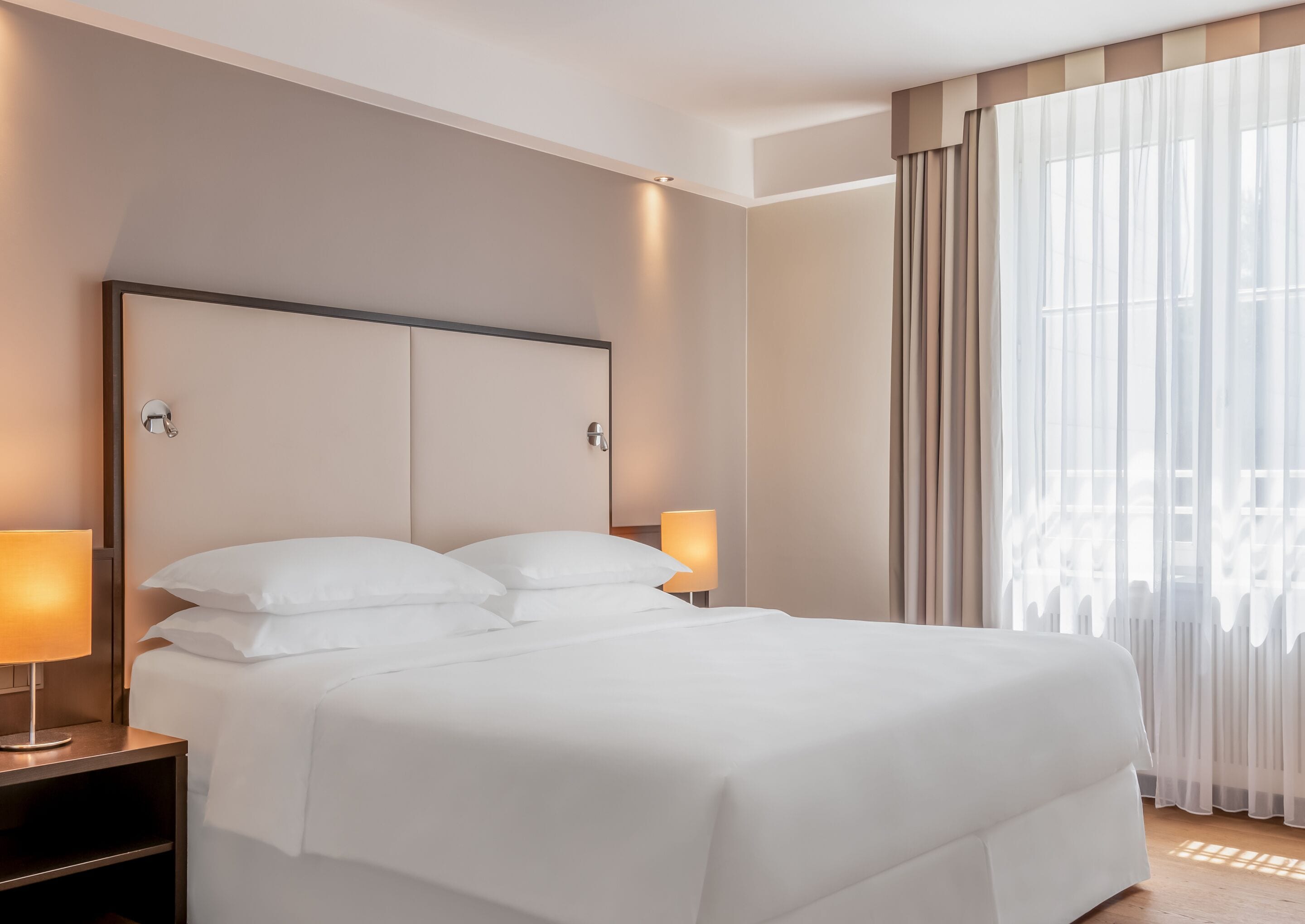 A modern hotel room with a large bed, two bedside lamps, and sheer curtains.