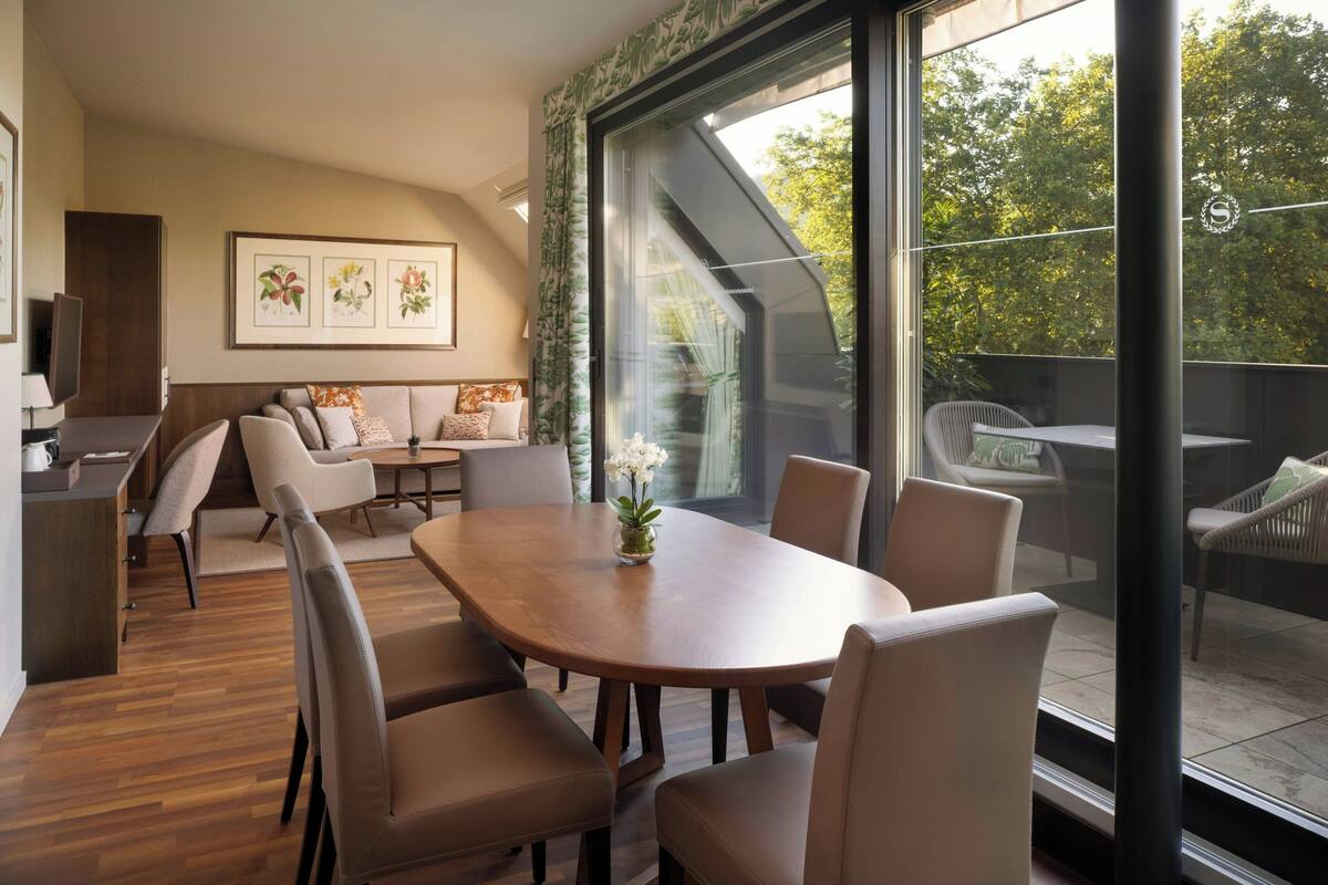Un comedor moderno con una mesa y sillas de madera, un florero y vistas al exterior a través de amplios ventanales.