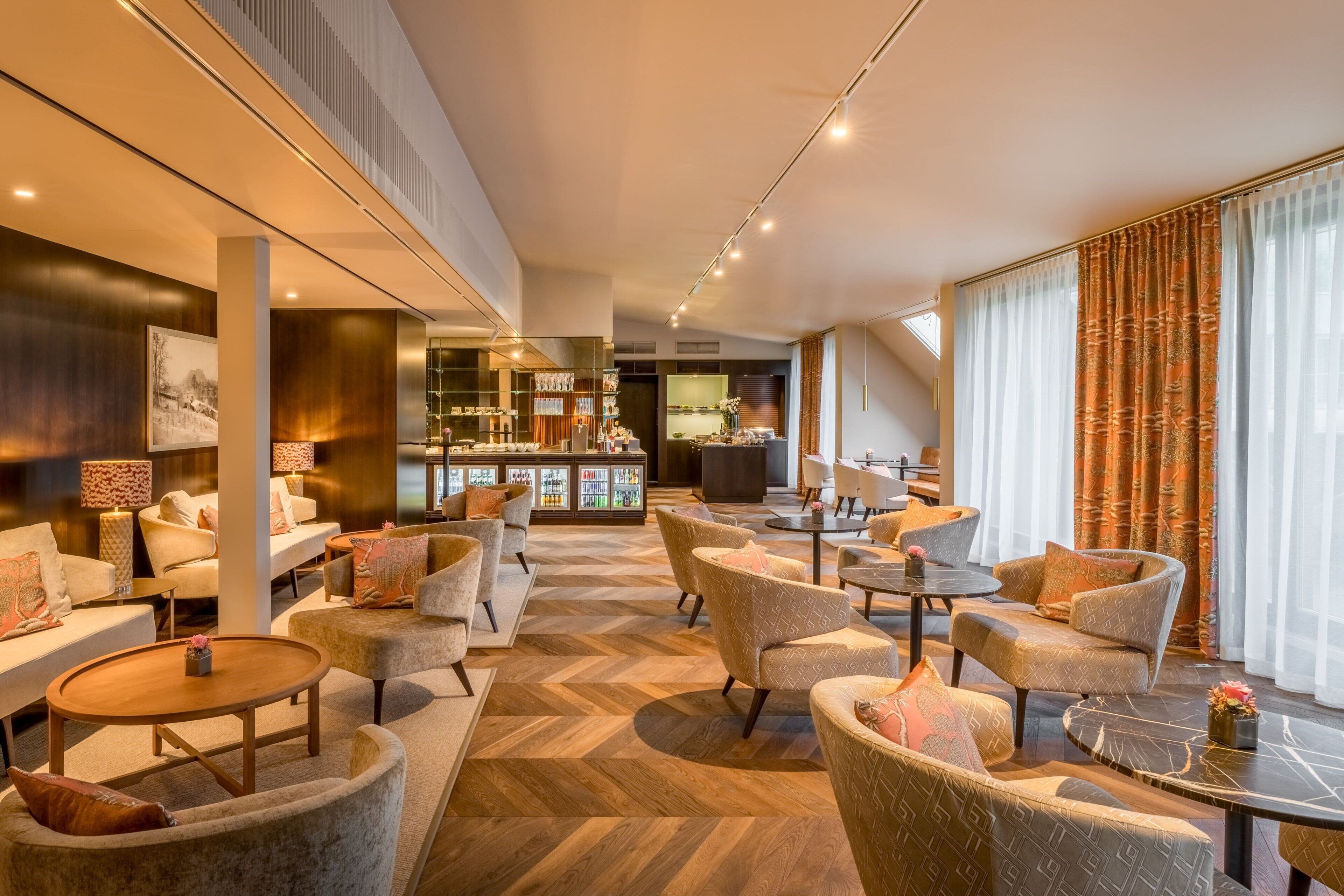 Eine geräumige Hotelhalle mit einem Fischgrätparkett, gemütlichen Sitzbereichen, einer Bar und großen Fenstern mit Vorhängen.