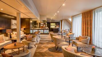 Eine geräumige Hotelhalle mit einem Fischgrätparkett, gemütlichen Sitzbereichen, einer Bar und großen Fenstern mit Vorhängen.