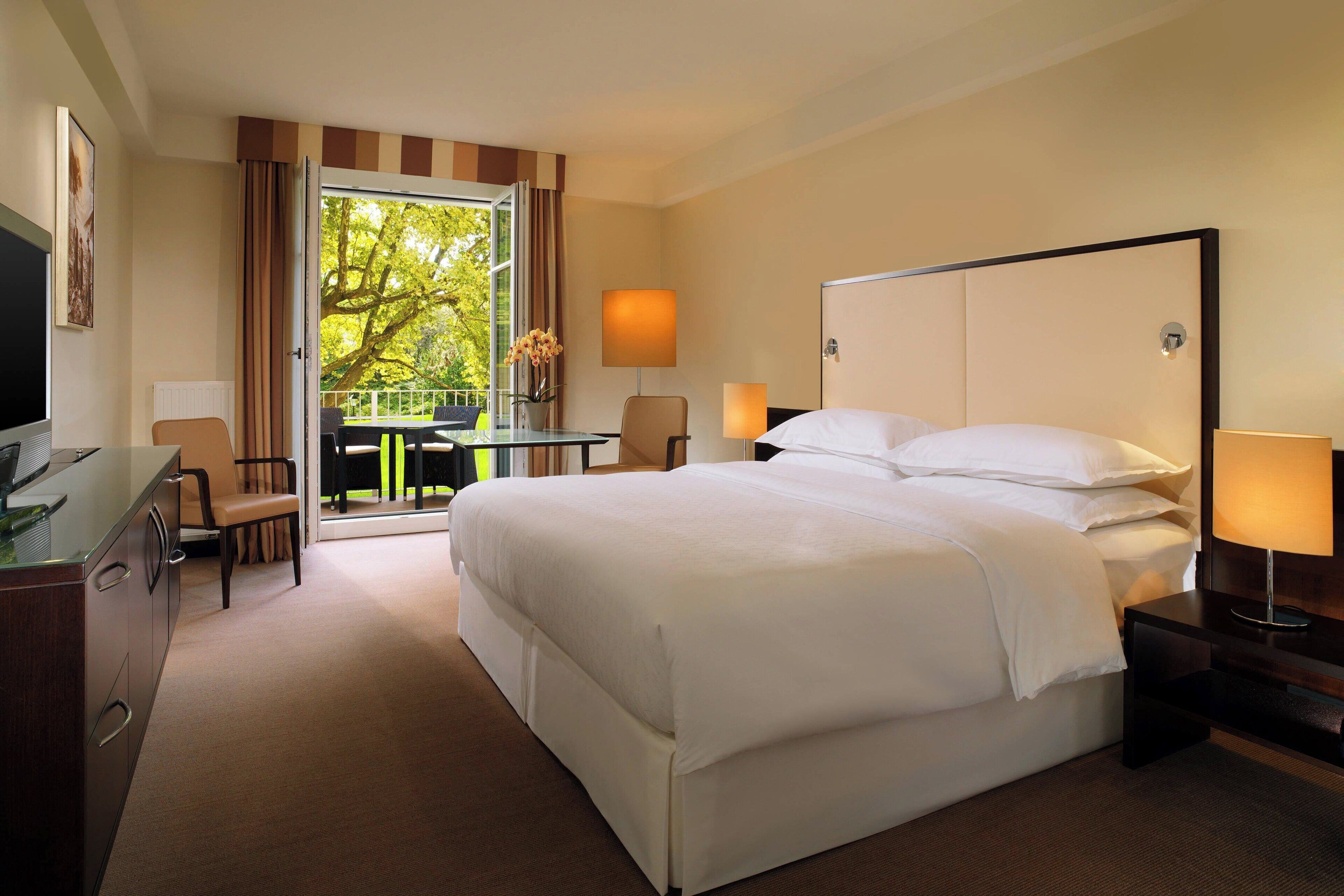 A hotel room with a large bed, a desk with a chair, a TV, and a balcony with a view of trees.