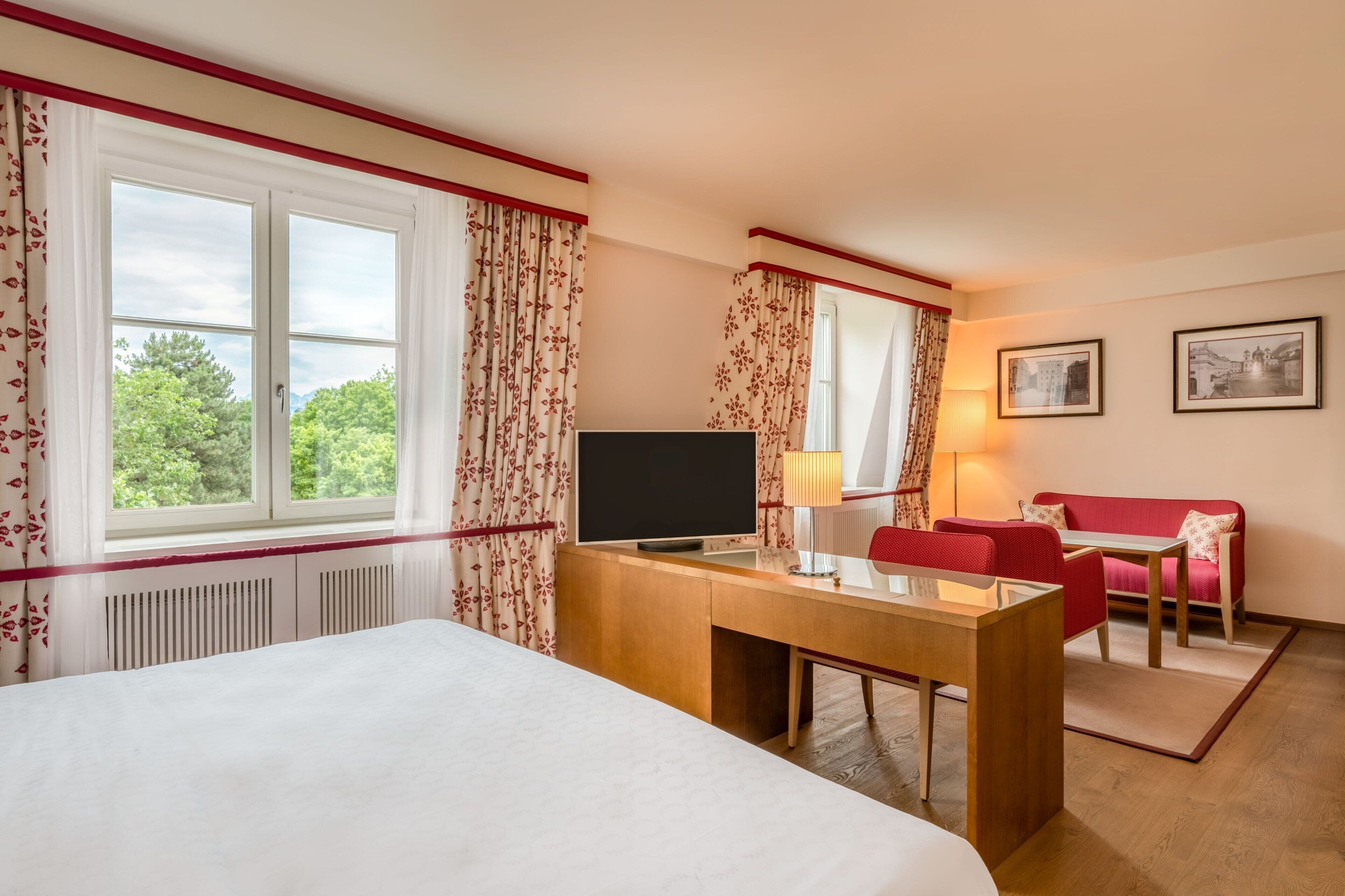 A hotel room with a bed, a desk, a TV, a red sofa, and a window with curtains.