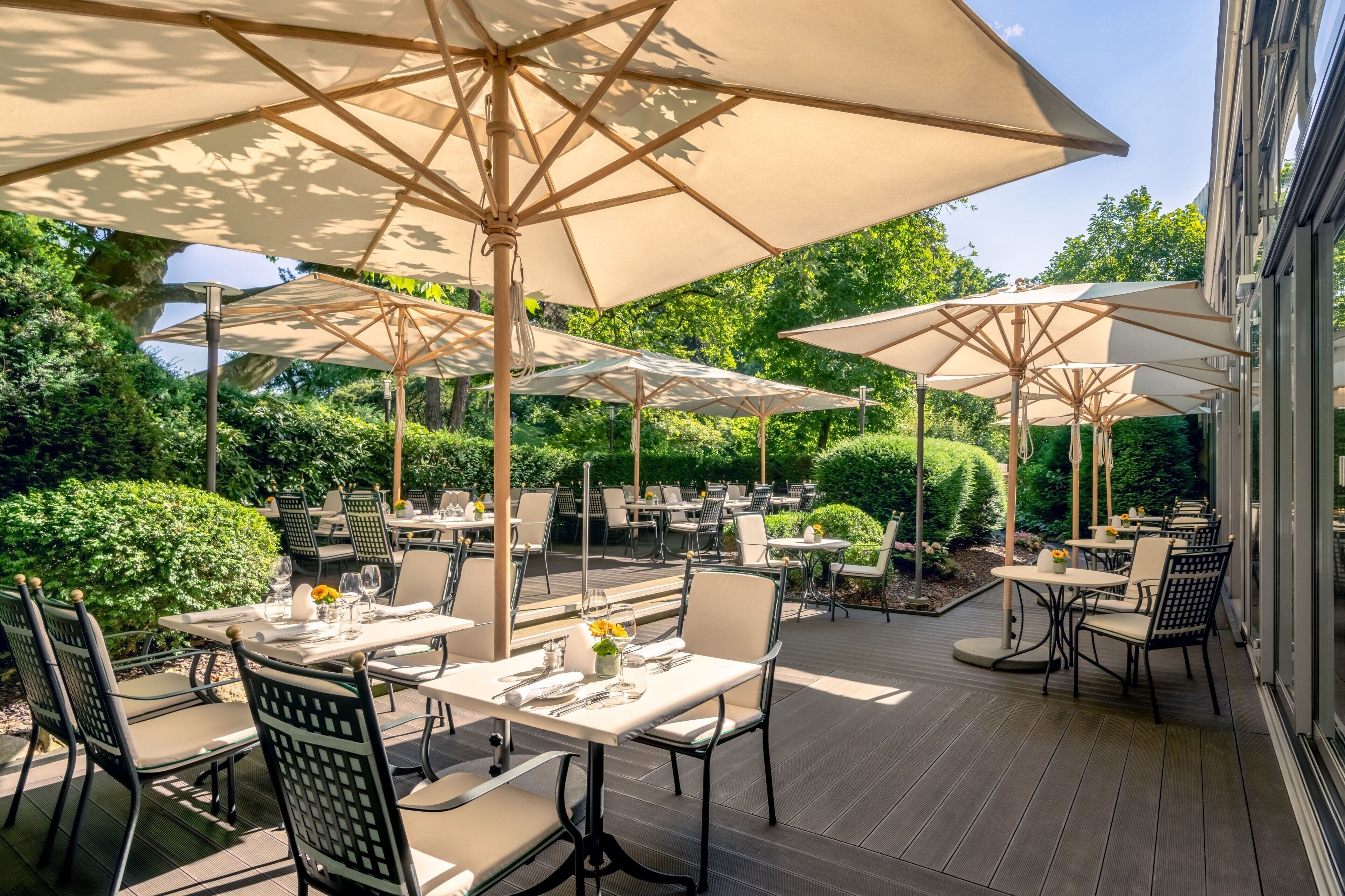 An outdoor patio with tables and chairs, large umbrellas, and greenery in the background.