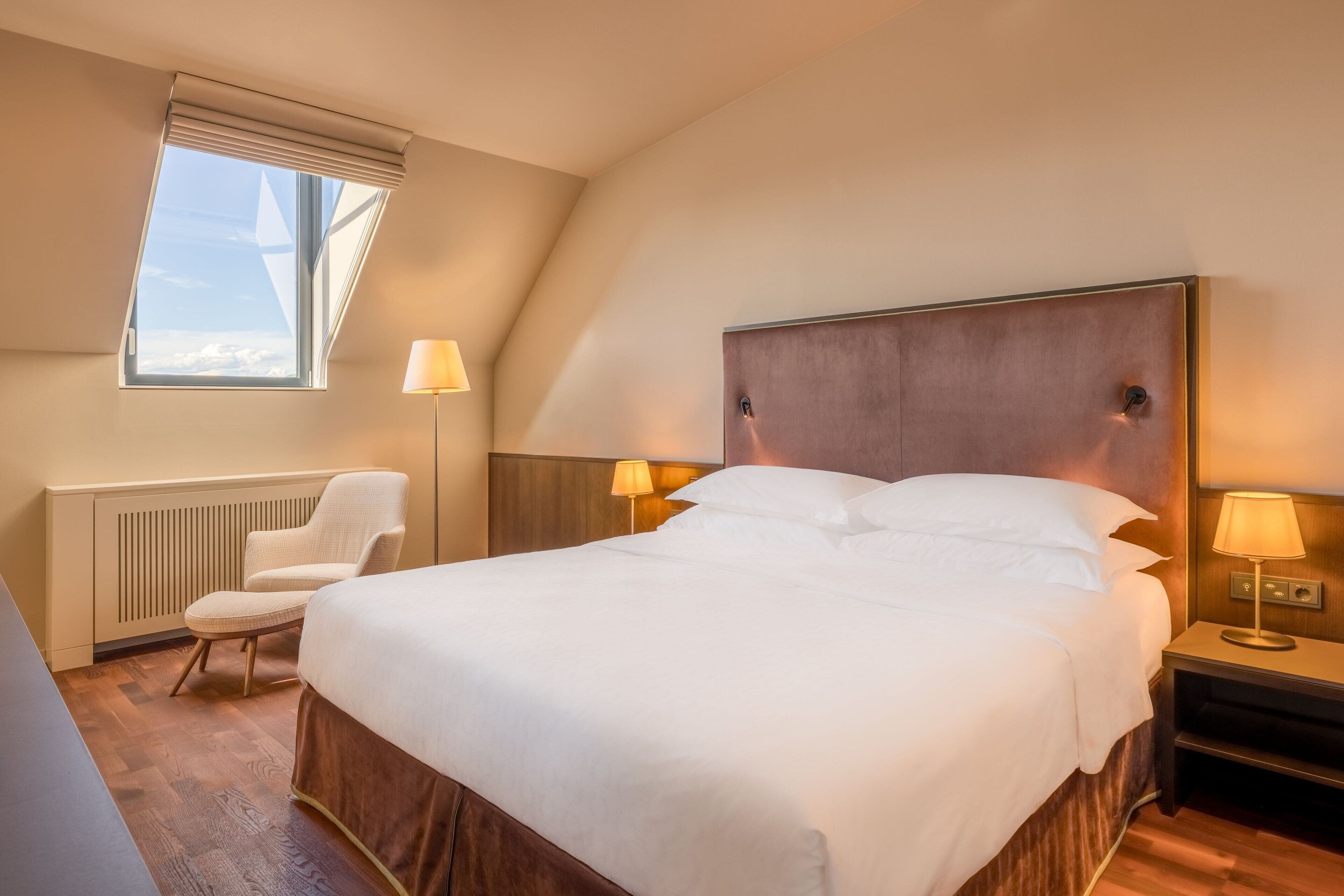 A hotel room with a large bed, a chair, a nightstand with lamps, a window with a view, and a radiator.