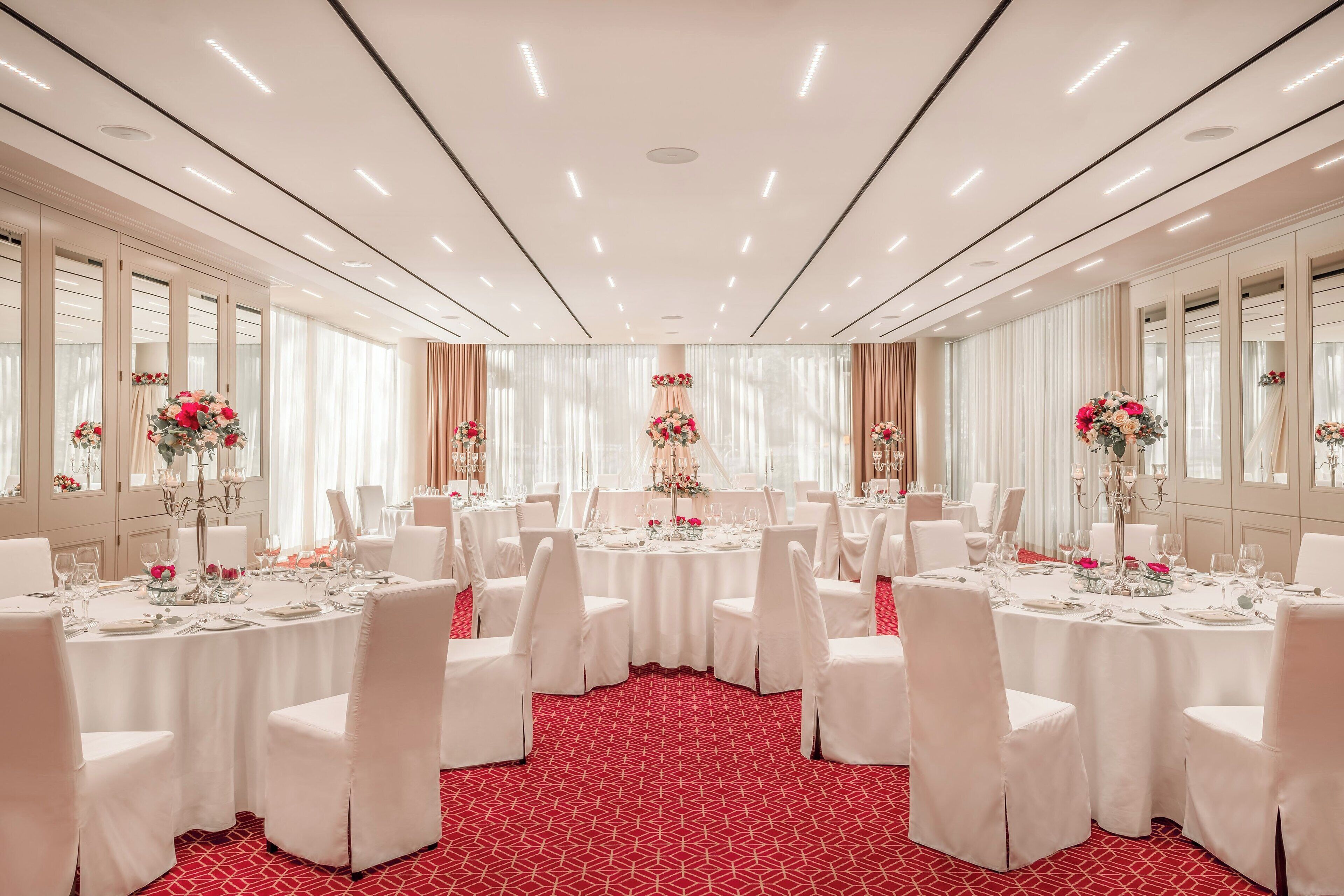 Un salón de banquetes con mesas redondas, sillas blancas y centros de mesa con flores.
