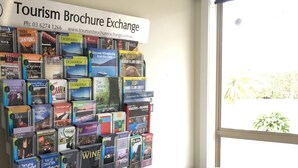 A wall with a Tourism Brochure Exchange and a grey armchair in a room with a window. - Seasonal South Motel & Function Centre (Ulverstone)