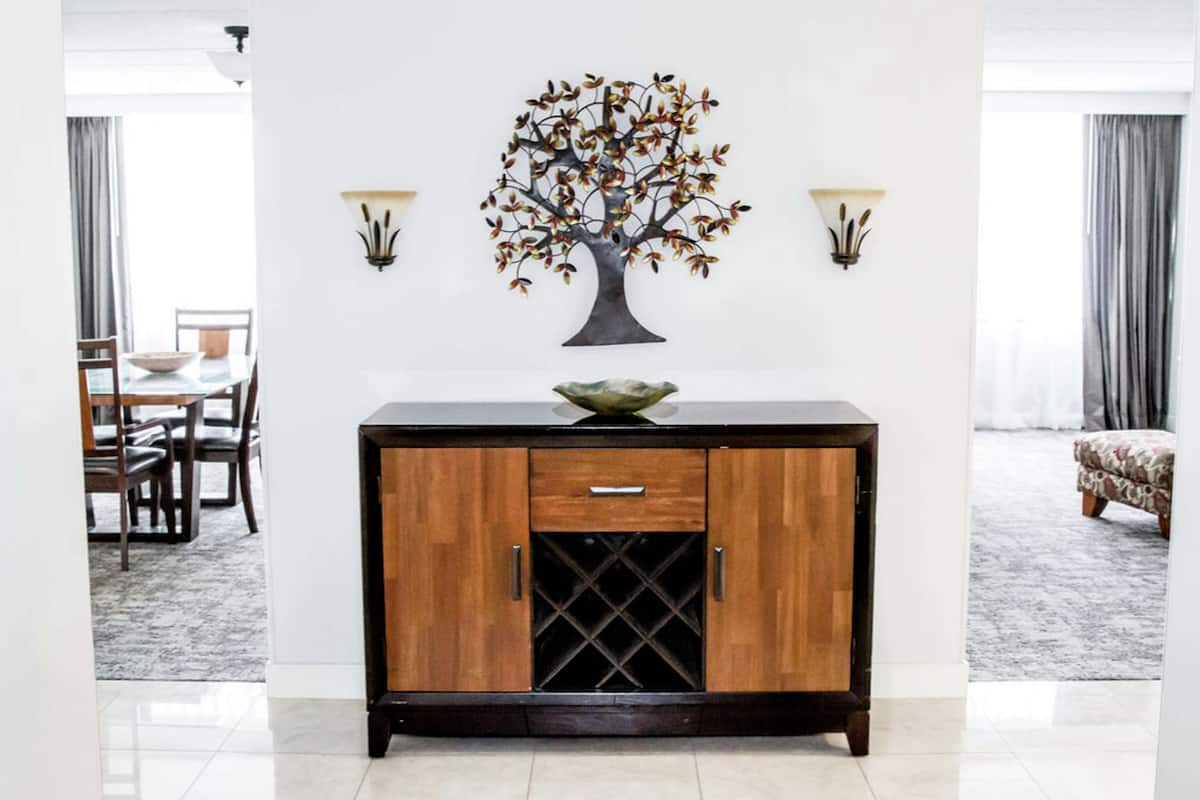 Un mueble de madera con un botellero, una planta decorativa y dos apliques de pared en una habitación desde la cual se ve un comedor a través de una puerta abierta.