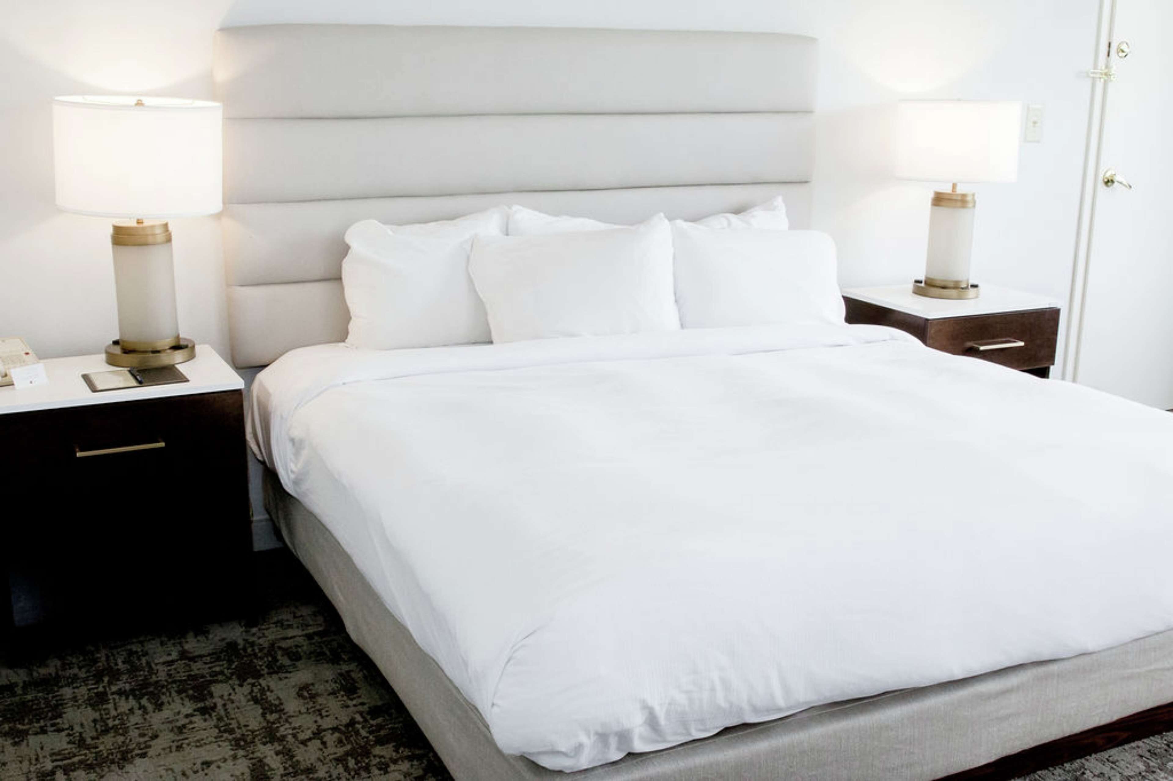 Una cama bien hecha con sábanas blancas, dos mesitas de noche con lámparas y una alfombra oscura en el suelo.