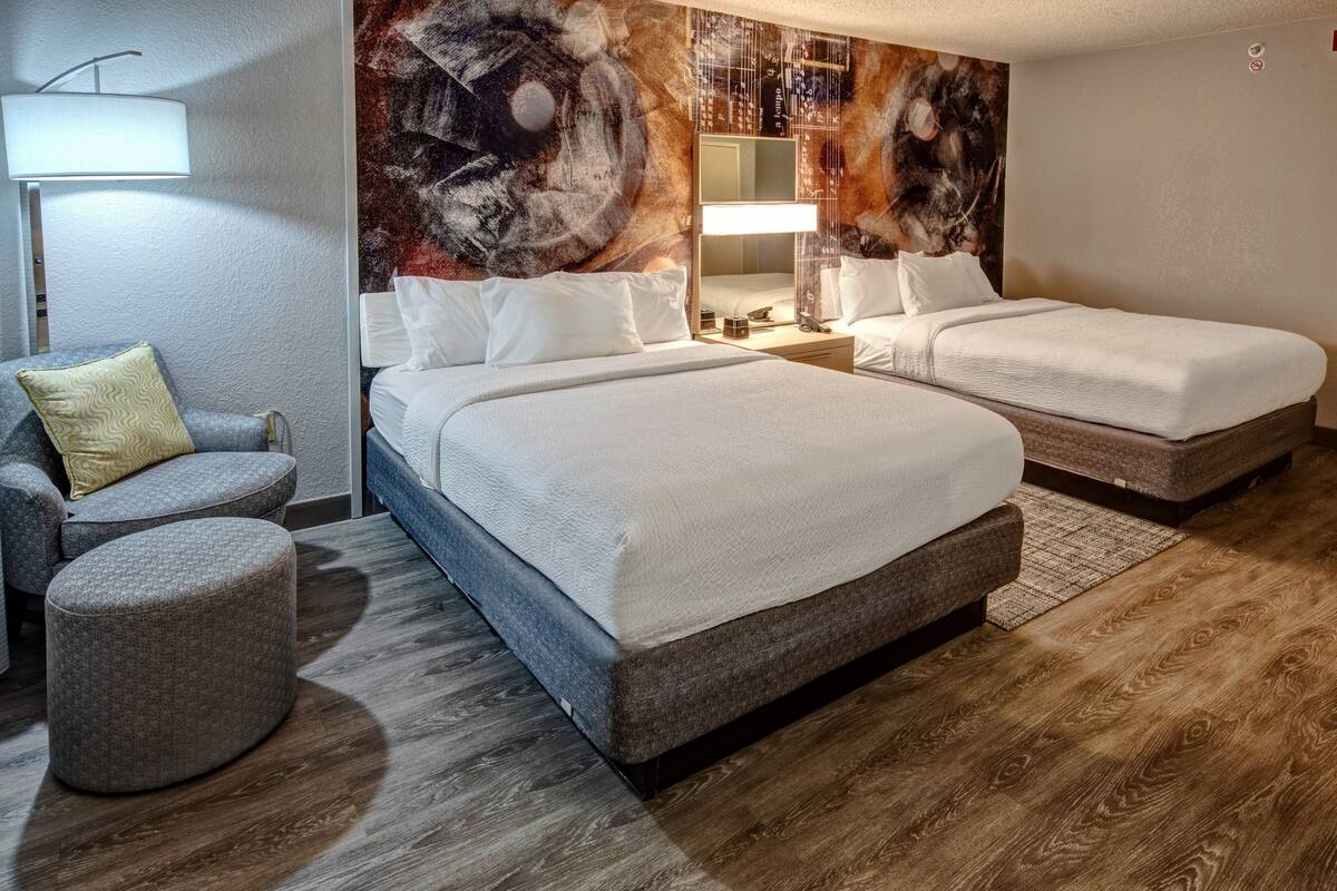 A hotel room with two beds, a seating area with a chair and ottoman, and a large abstract painting on the wall.