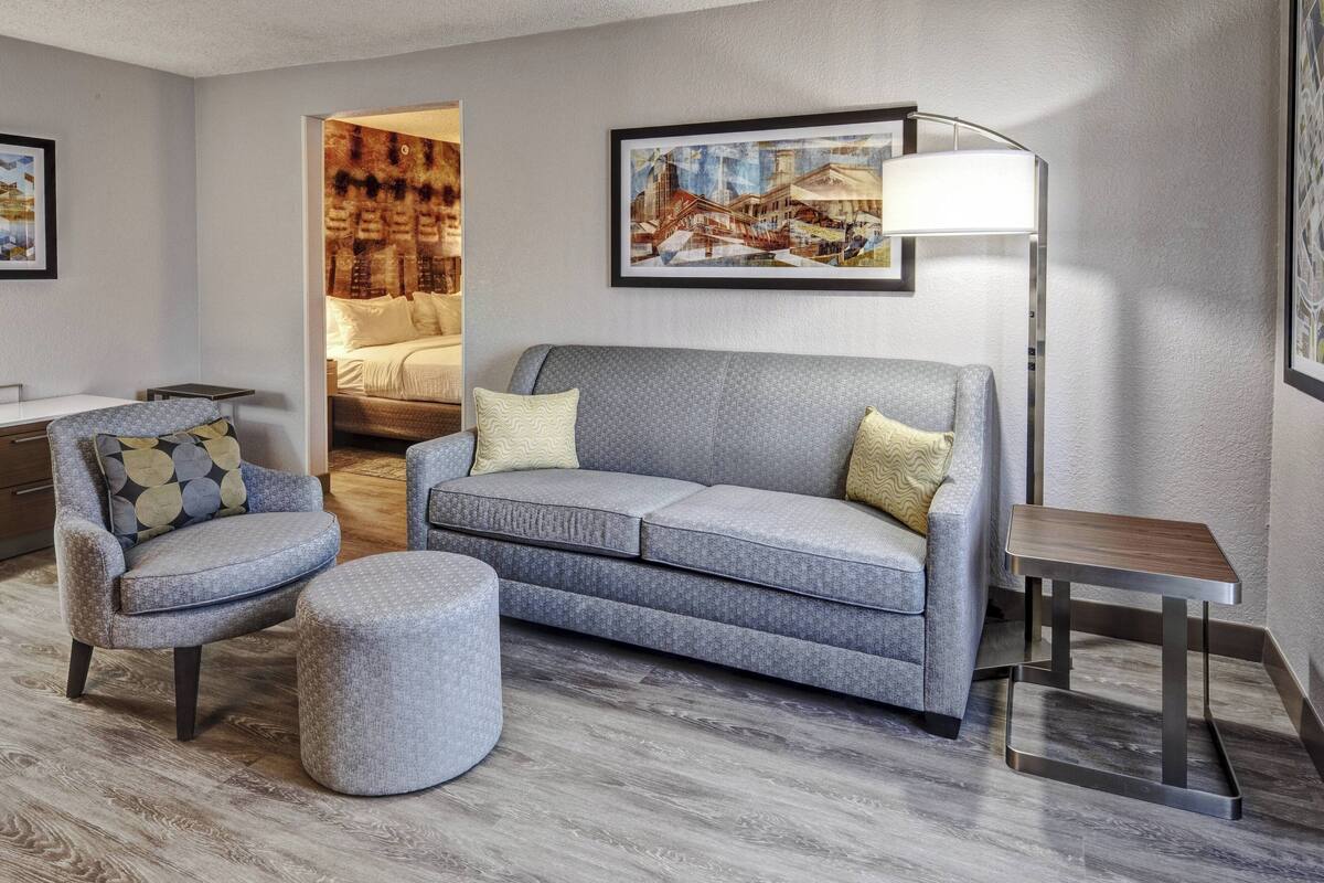 A modern living room with a grey sofa, matching ottoman, and a small table. There is a painting on the wall and a lamp beside the sofa.