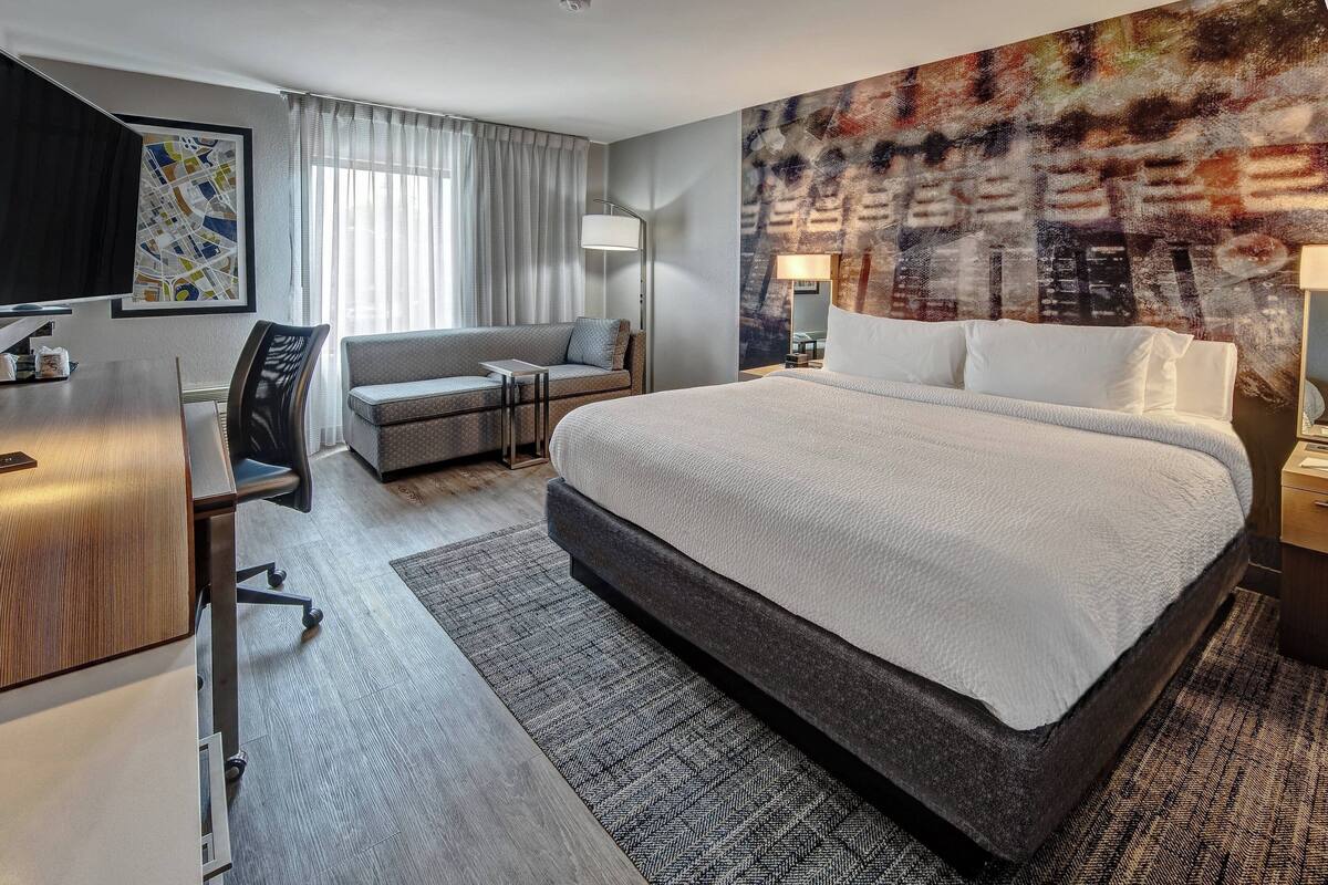 A modern hotel room with a large bed, a desk, a sofa, and a large abstract painting on the wall.