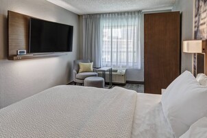 A hotel room with a large bed, a flat-screen TV mounted on the wall, a small table with a chair, and a window with curtains.