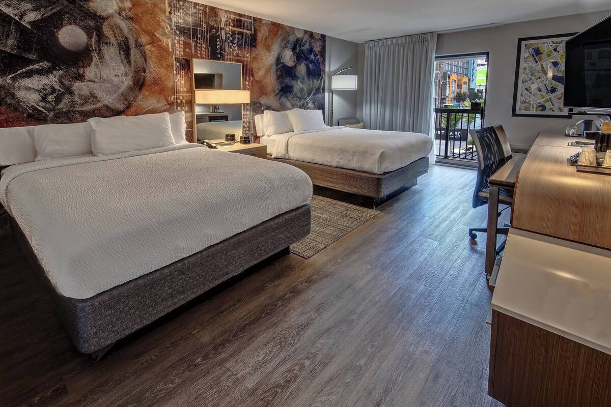 A hotel room with two beds, a desk, a chair, a TV, and a large window with a view of the city.
