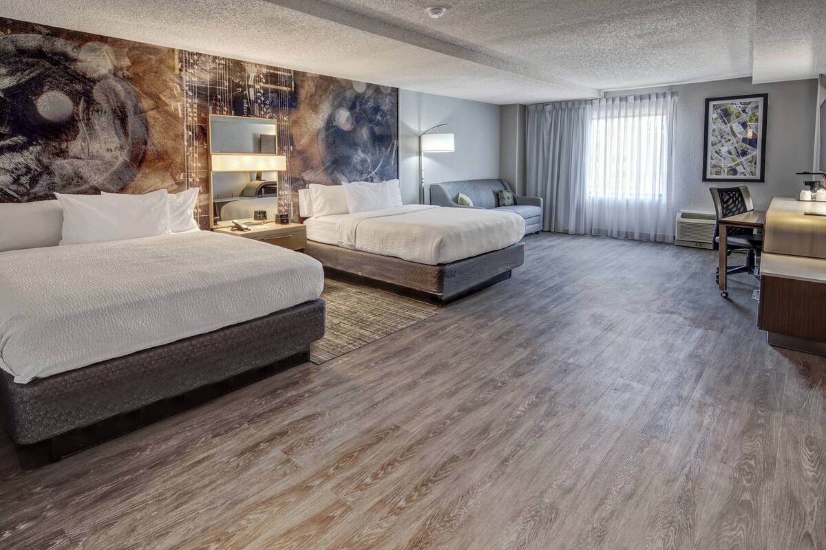 A hotel room with two beds, a desk, a chair, a large abstract painting, and a window with curtains.