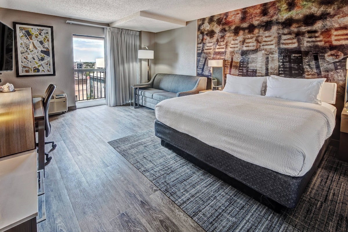A hotel room with a large bed, a sofa, a desk, and a view of the city through the balcony.
