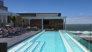 A rooftop infinity pool with a glass railing overlooking the ocean, lounge chairs, and a wooden deck. - The Blue Waters Hotel (Durban)
