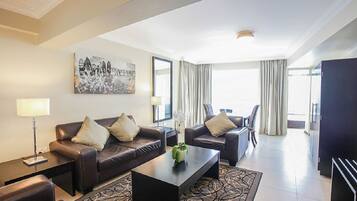 A modern hotel room with a brown leather sofa, a matching armchair, a coffee table, and a large window with curtains.