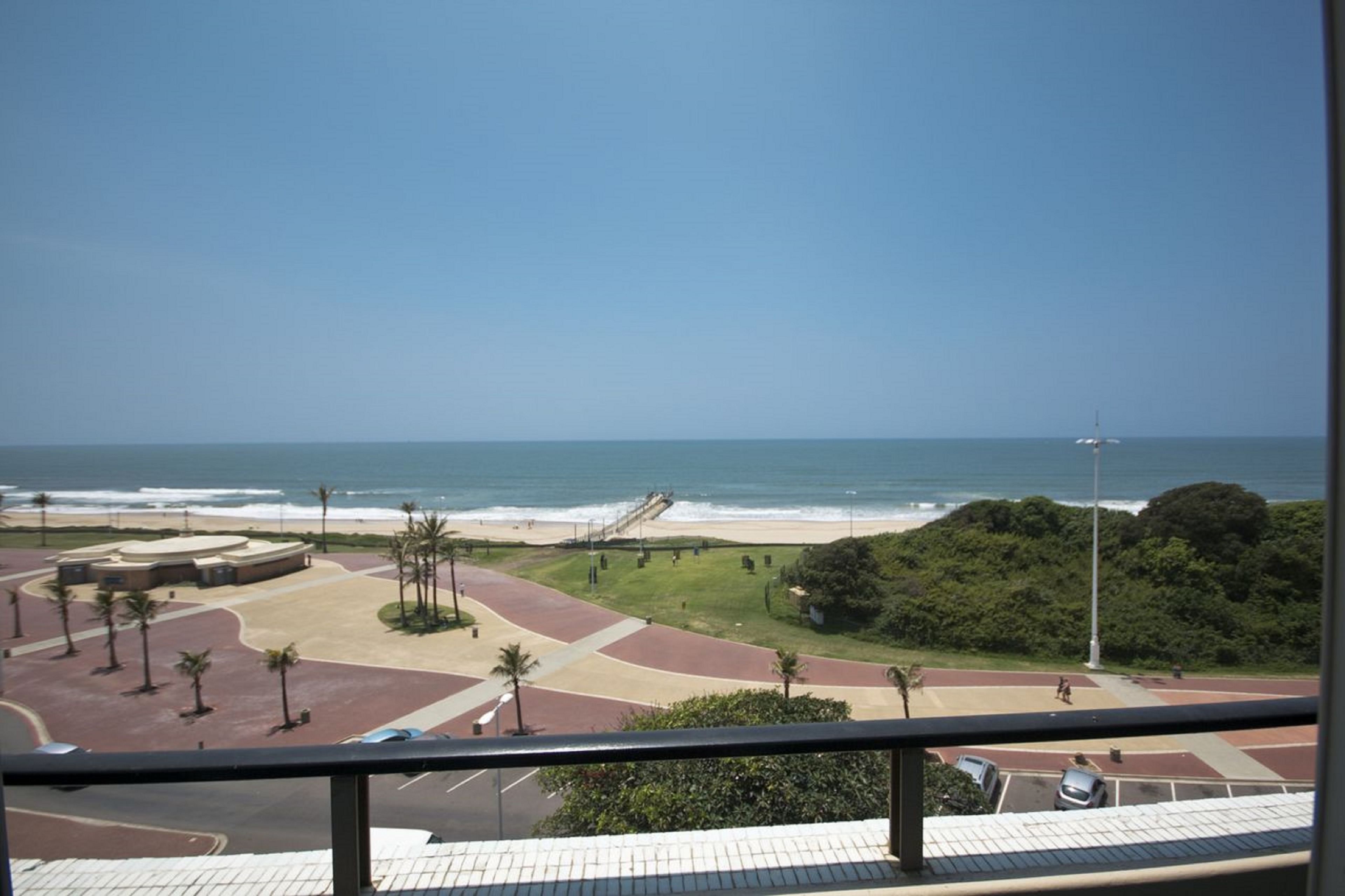 A coastal view from a balcony with a beach, palm trees, and a pedestrian path.