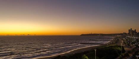 Un paisaje urbano costero al atardecer con una playa, paseo marítimo y edificios altos.