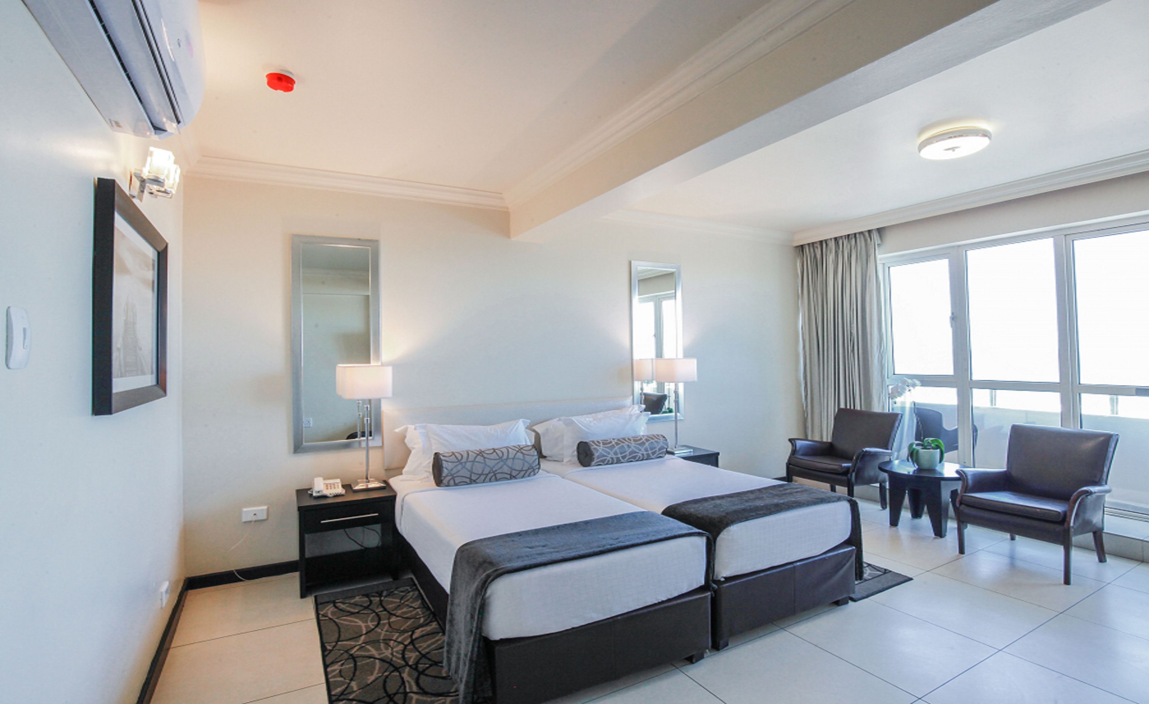 A hotel room with a double bed, two bedside tables, a desk with a chair, a large window with curtains, and a mirror on the wall.