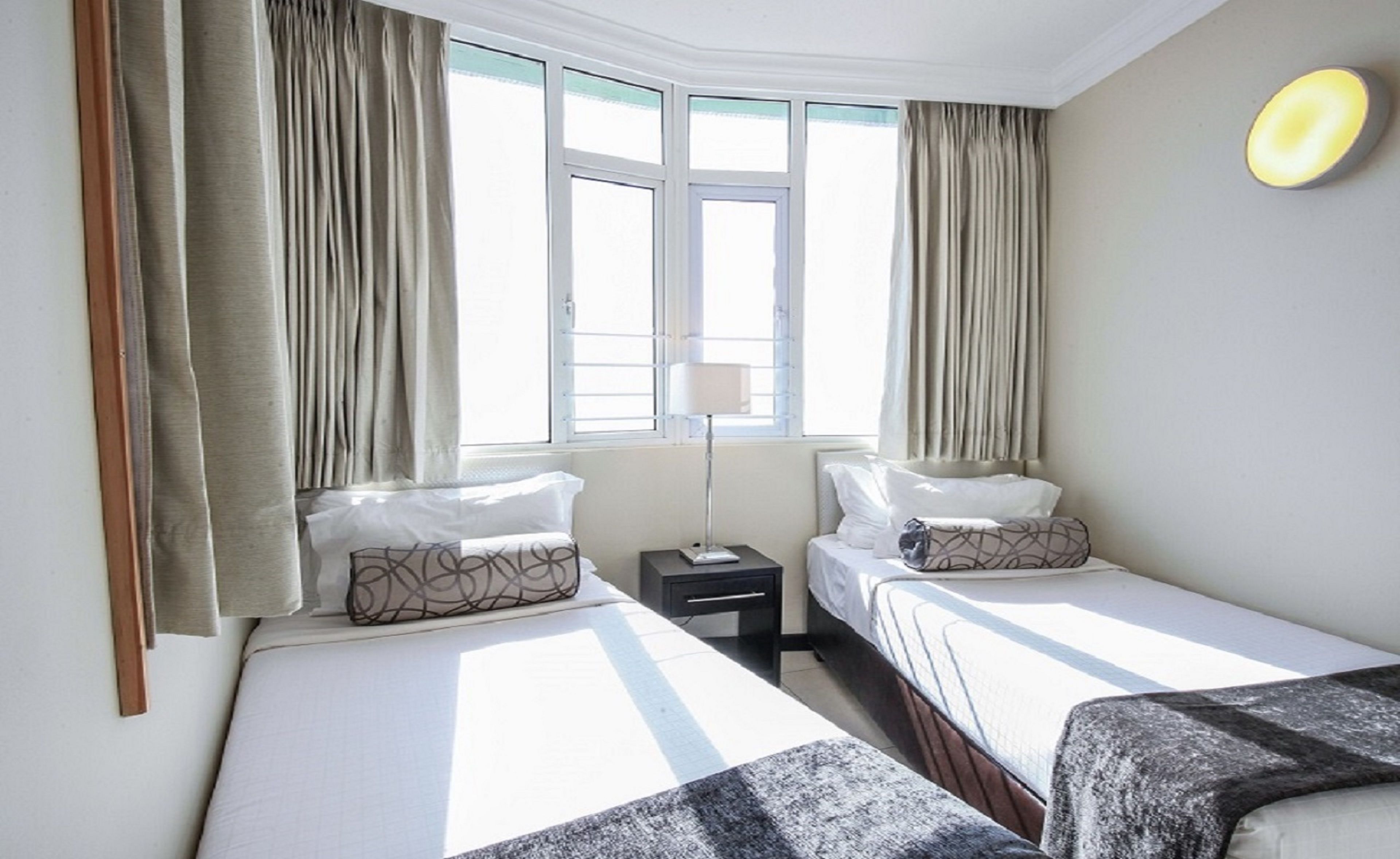 A hotel room with two beds, a nightstand, a lamp, and a window with curtains.