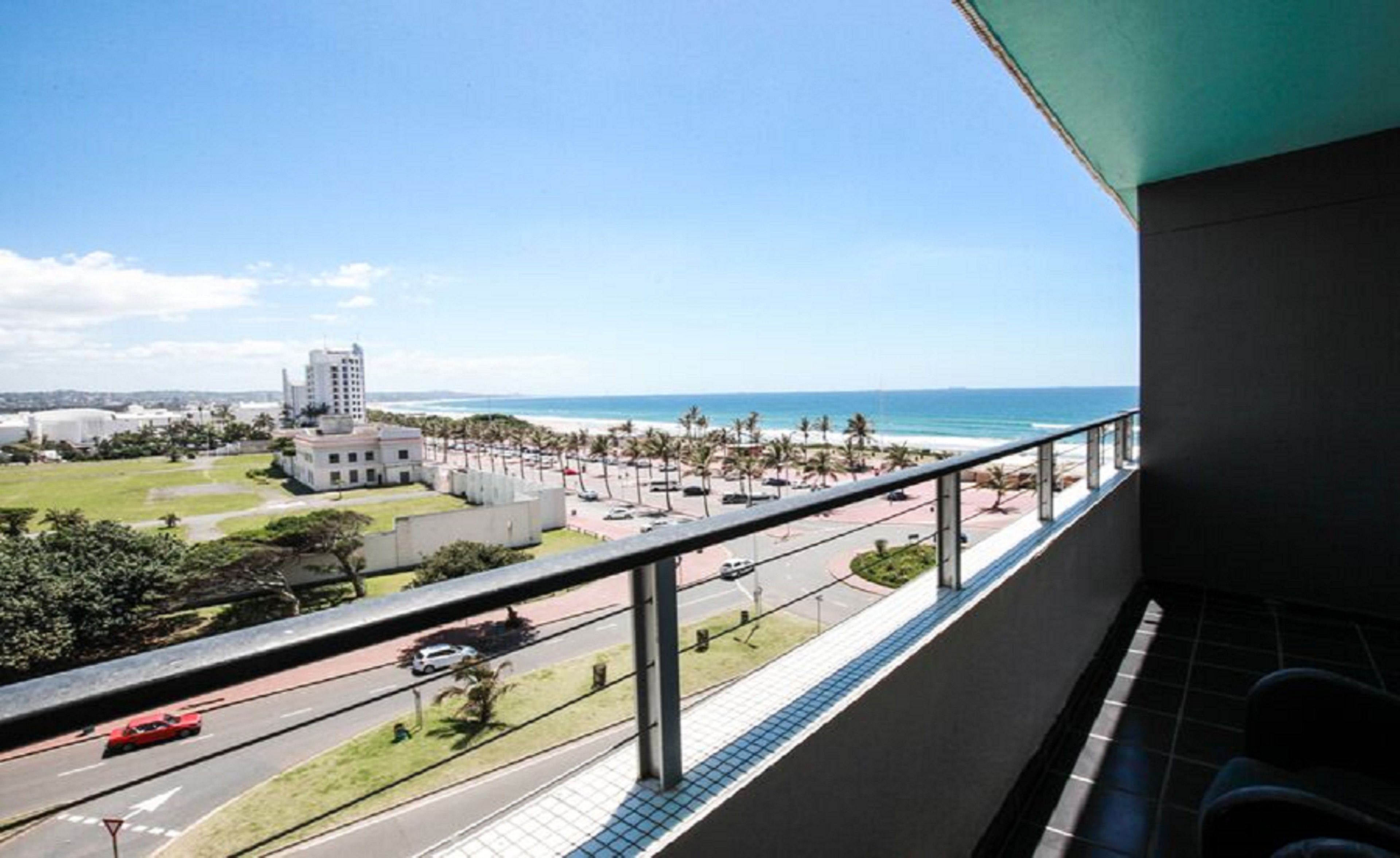 A balcony with a view of a beach, a road with cars, and buildings in the distance.