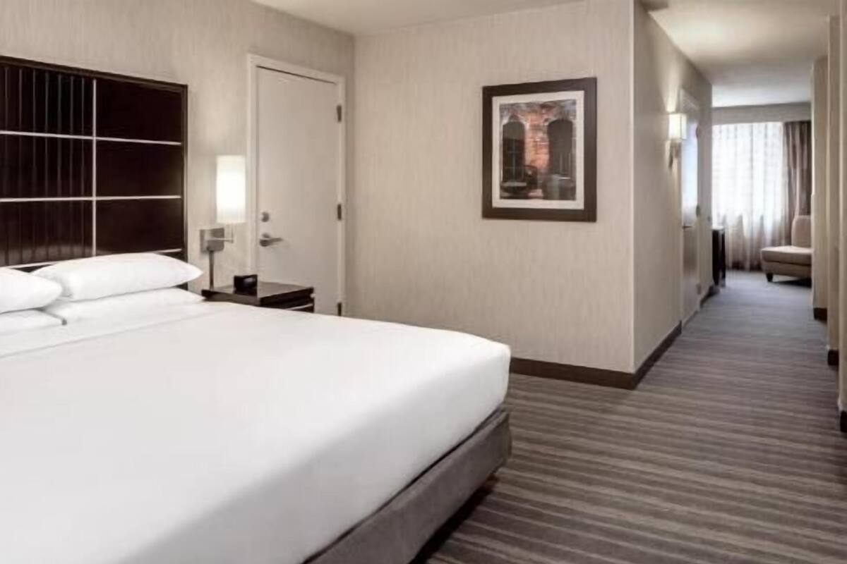 Hotellihuone, jossa on suuri sänky, yöpöytä, seinällä oleva taulu ja matolla peitetty käytävä.