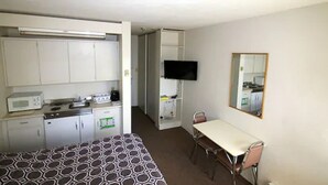 A compact hotel room with a kitchenette, a single bed, a dining table with chairs, a TV mounted on the wall, and a mirror.