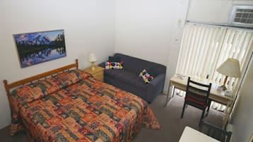 A small, neatly arranged room with a bed, a sofa, a desk, and a chair. There is a painting on the wall and a window with blinds.