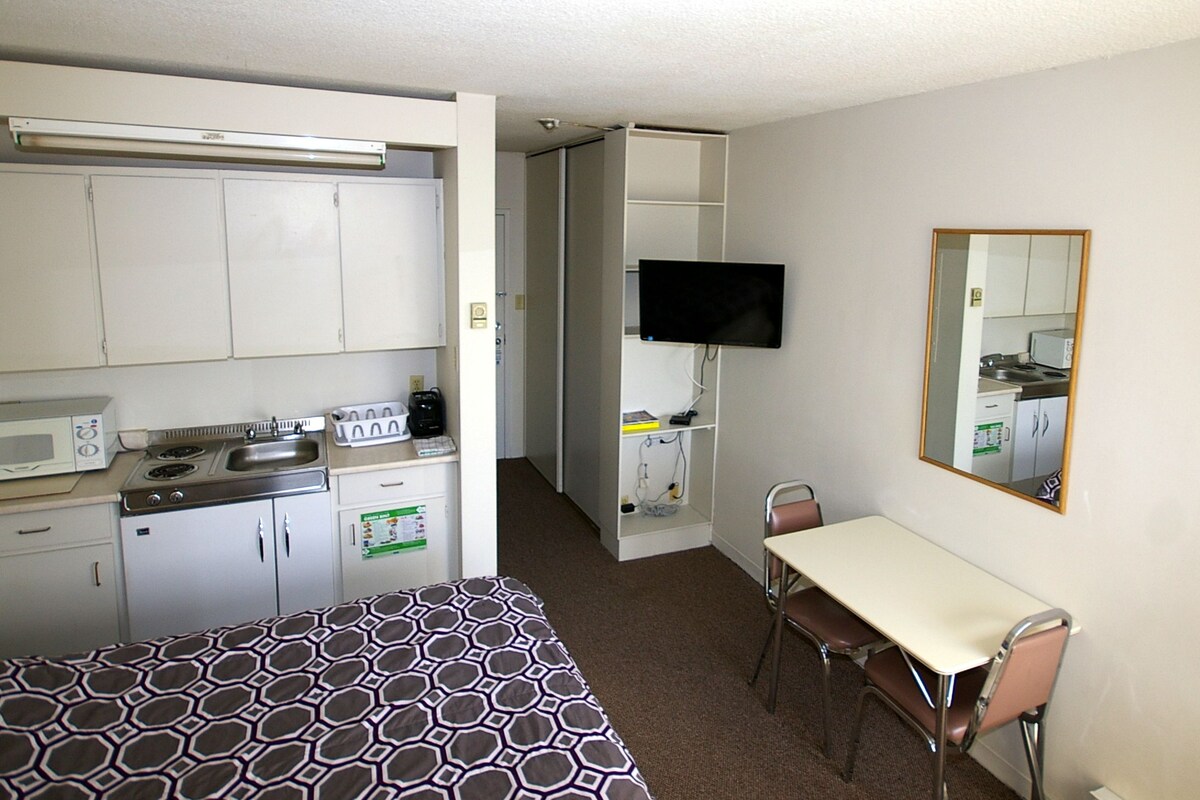 A compact hotel room with a kitchenette, a bed with a patterned bedspread, a desk with a chair, and a television mounted on the wall.