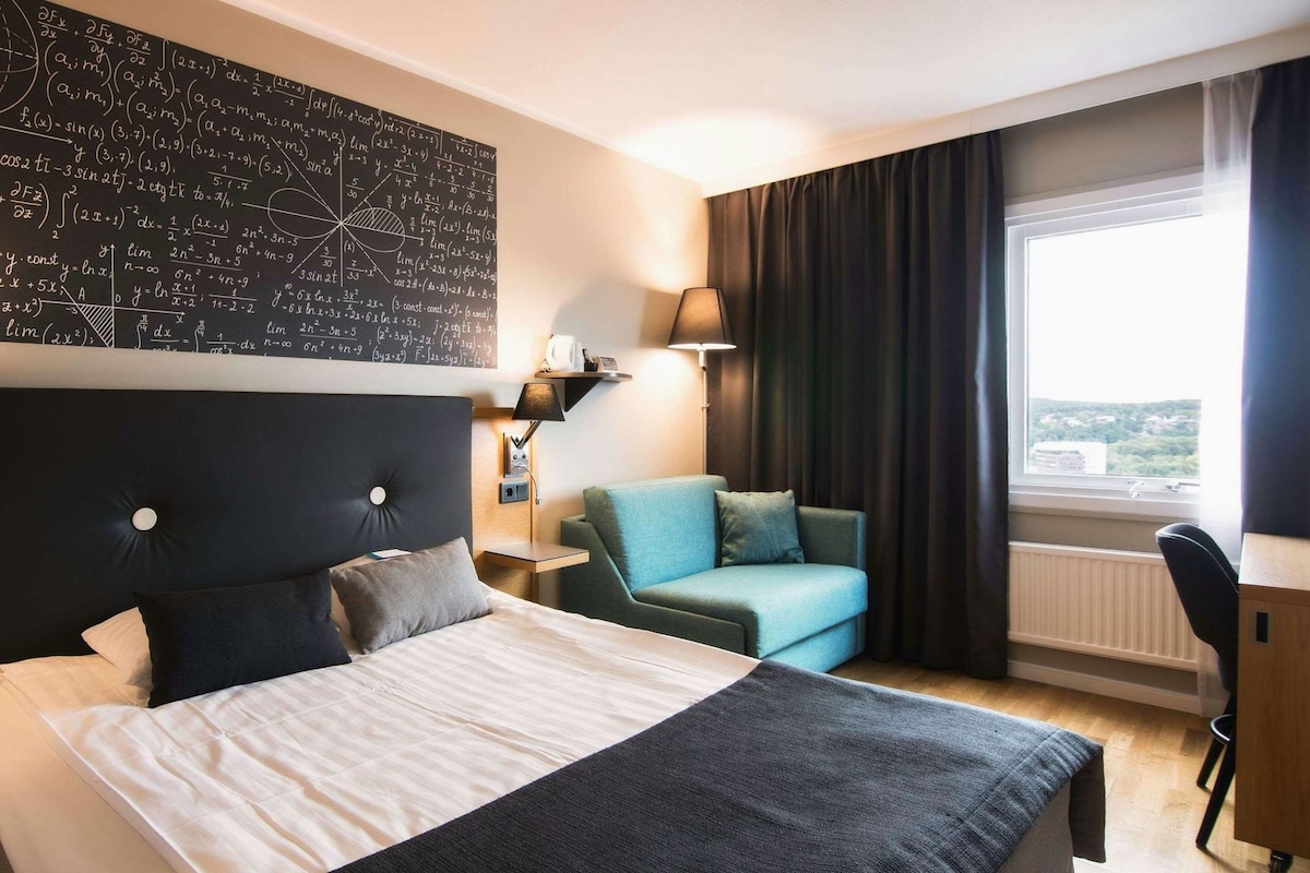 A hotel room with a bed, a desk, a chair, a window with curtains, and a wall with mathematical equations.