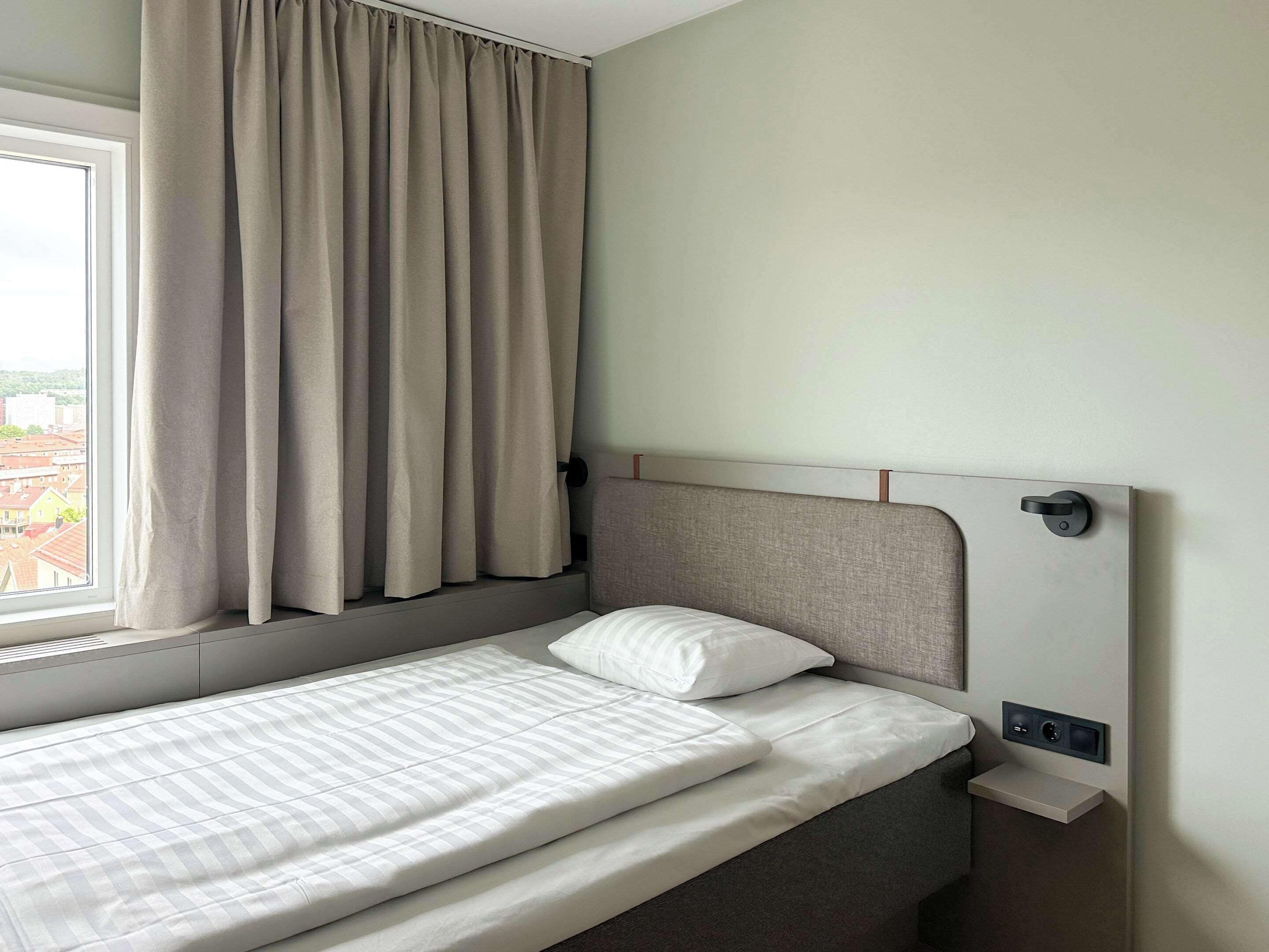 Een eenpersoonsbed met hoofdbord, een raam met gordijnen en een bedieningspaneel aan de muur.