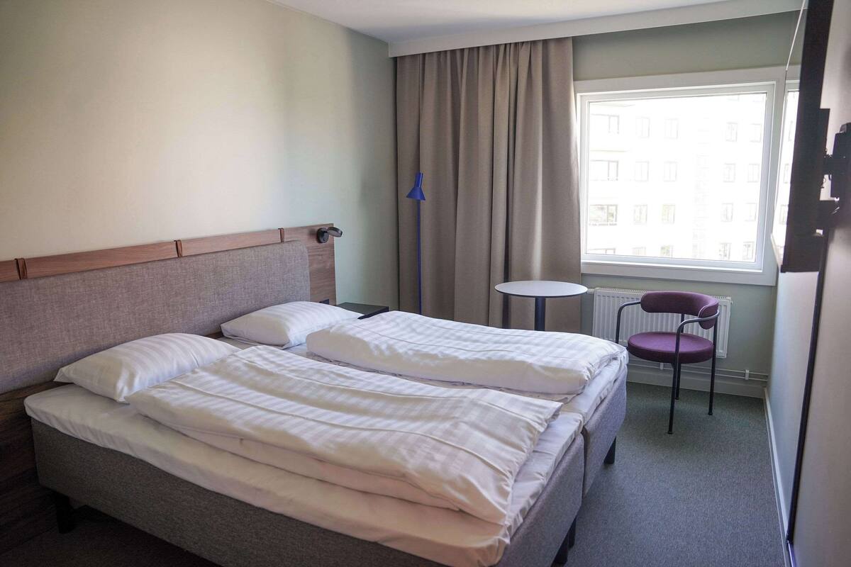 Quarto de hotel com cama, cadeira, mesinha e janela com cortinas.