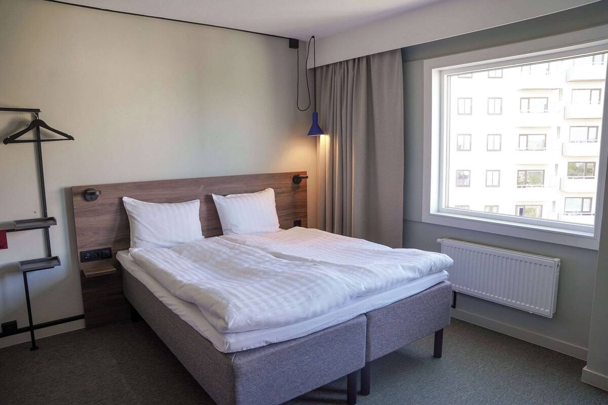 Uma cama bem arrumada com cabeceira cinza, uma mesa de cabeceira com abajur e uma janela com cortinas.