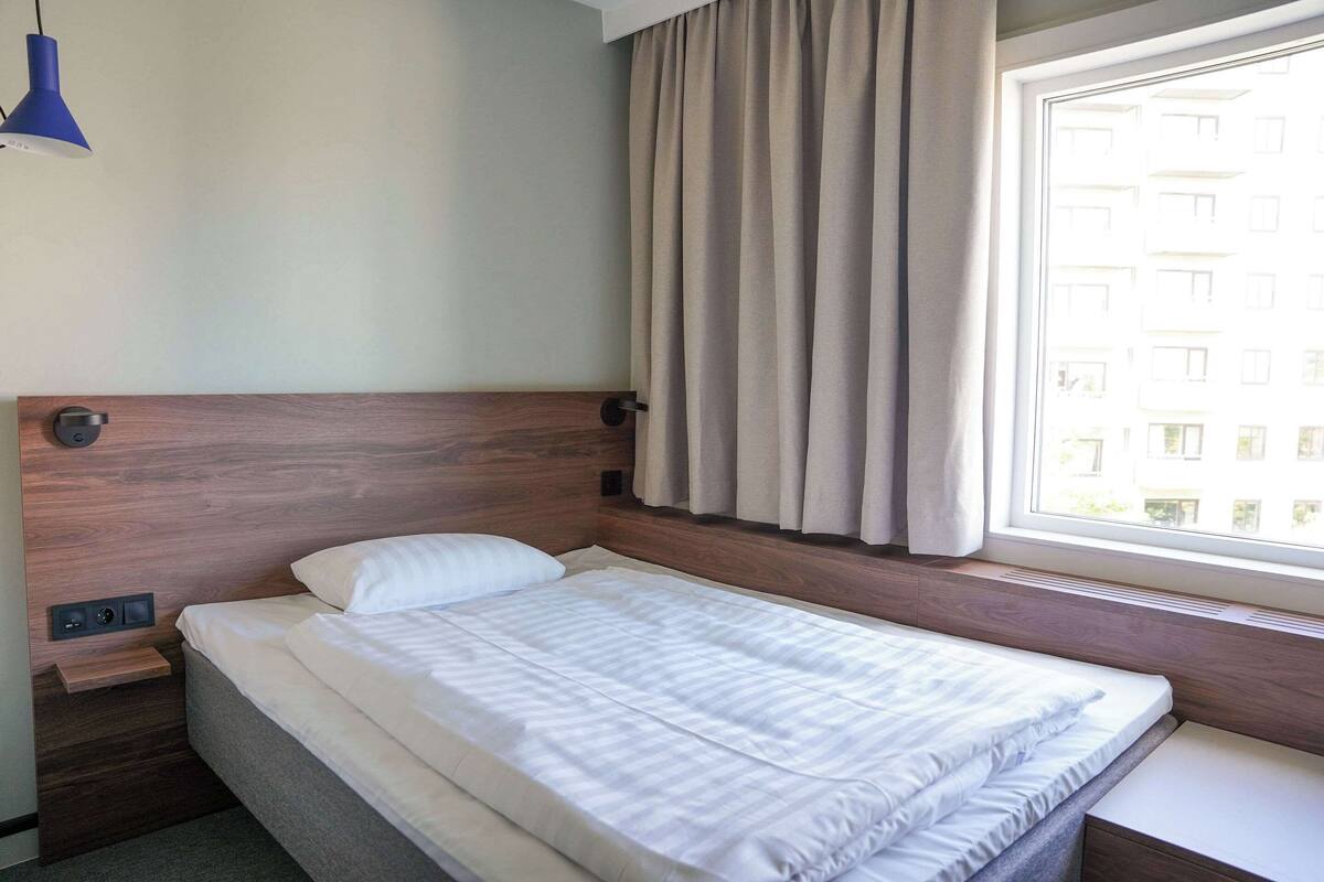 Uma cama bem arrumada com cabeceira de madeira, mesa de cabeceira com tomada e janela com cortinas.