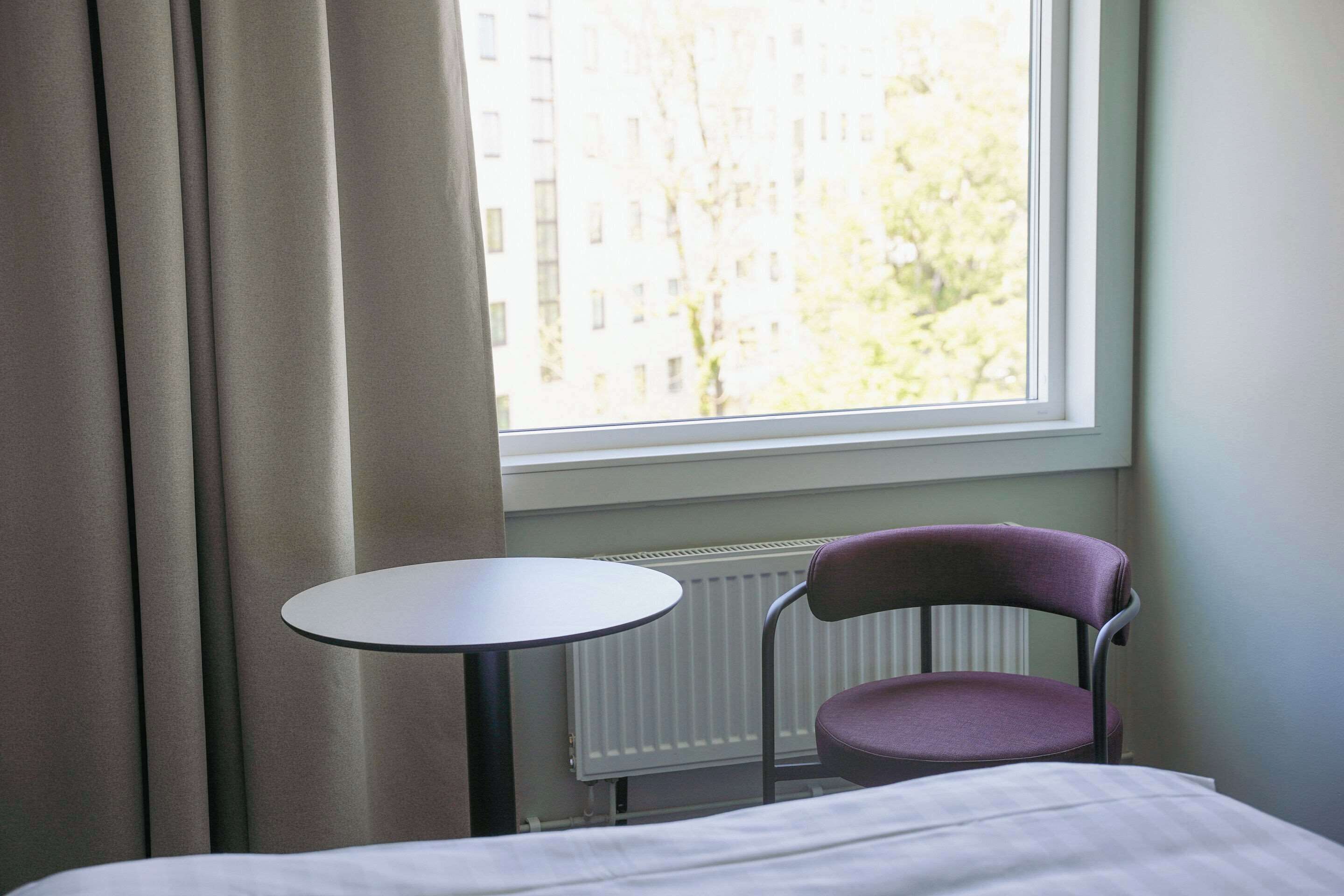 Ein Hotelzimmer mit einem Bett, einem kleinen runden Tisch, einem violetten Sessel und einem Fenster mit Vorhängen.
