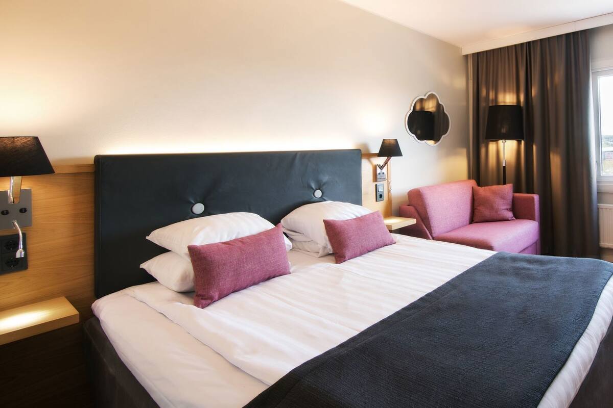Quarto de hotel com uma cama, duas cadeiras, uma escrivaninha, um abajur e vista para o exterior.