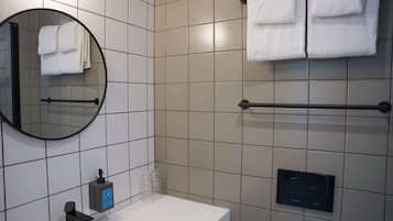 Un bany amb un mirall rodó, un lavabo blanc, un suport de tovalloles amb tovalloles doblegades, una barra de tovalloles i un dosificador de sabó.