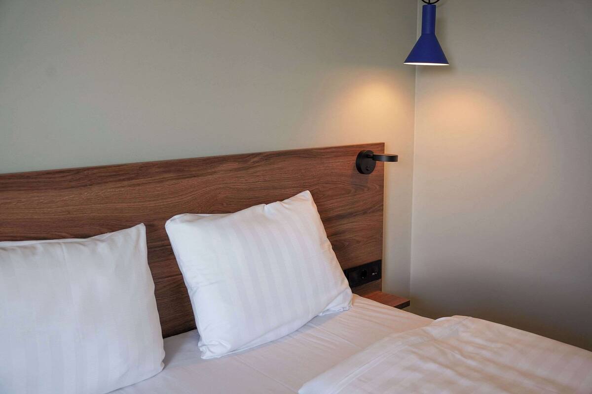Cama bem arrumada com lençóis brancos e duas almofadas, cabeceira de madeira e luminária azul fixada na parede.