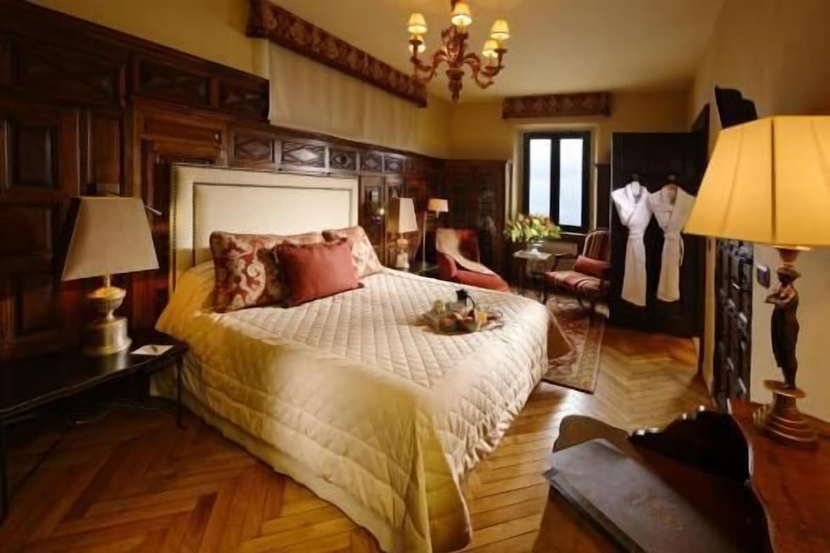 A spacious bedroom with a large bed, wooden paneling, a chandelier, and a sitting area with a chair and table.