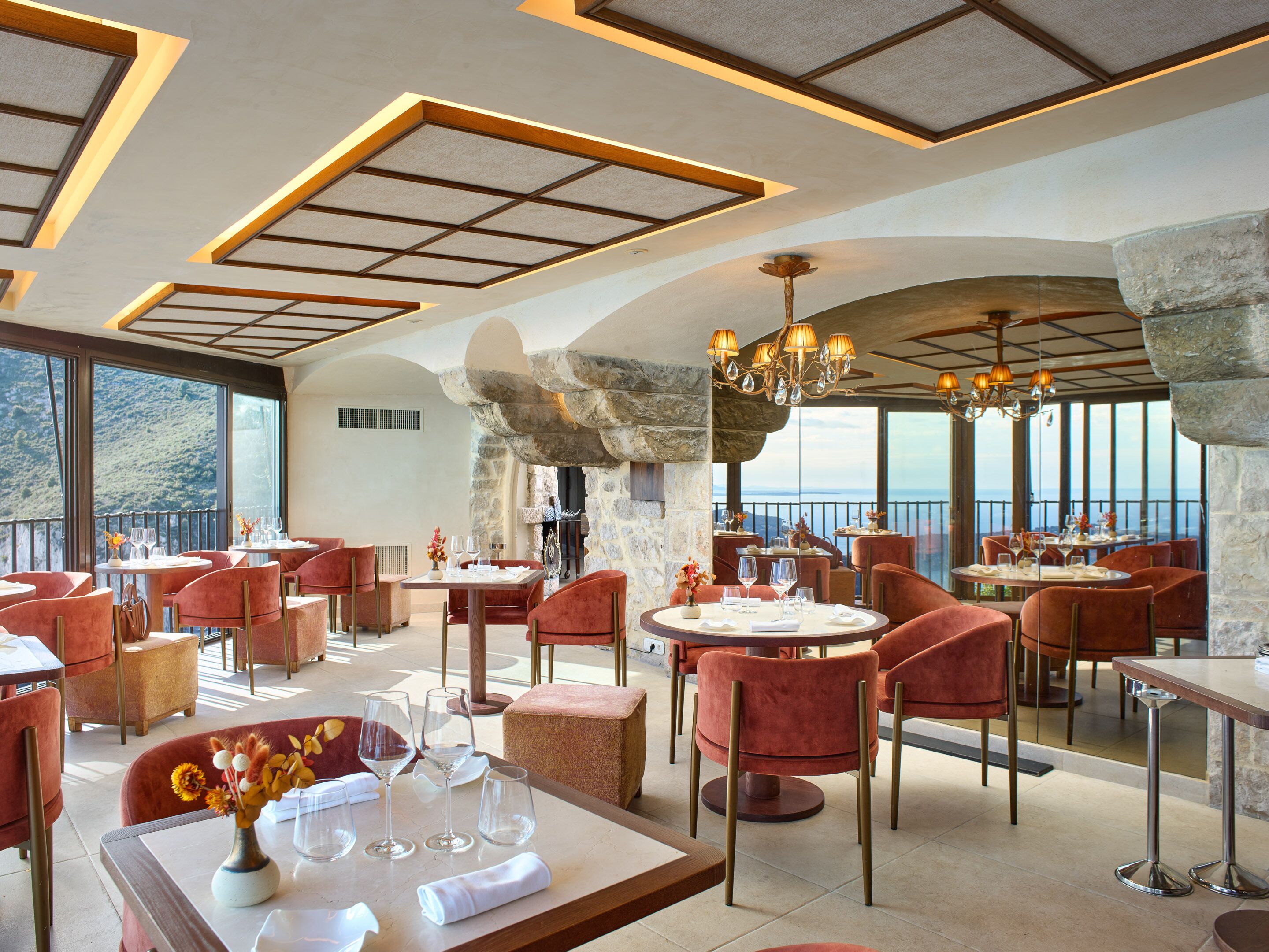 A restaurant with tables and chairs, a chandelier, and a view of the sea.