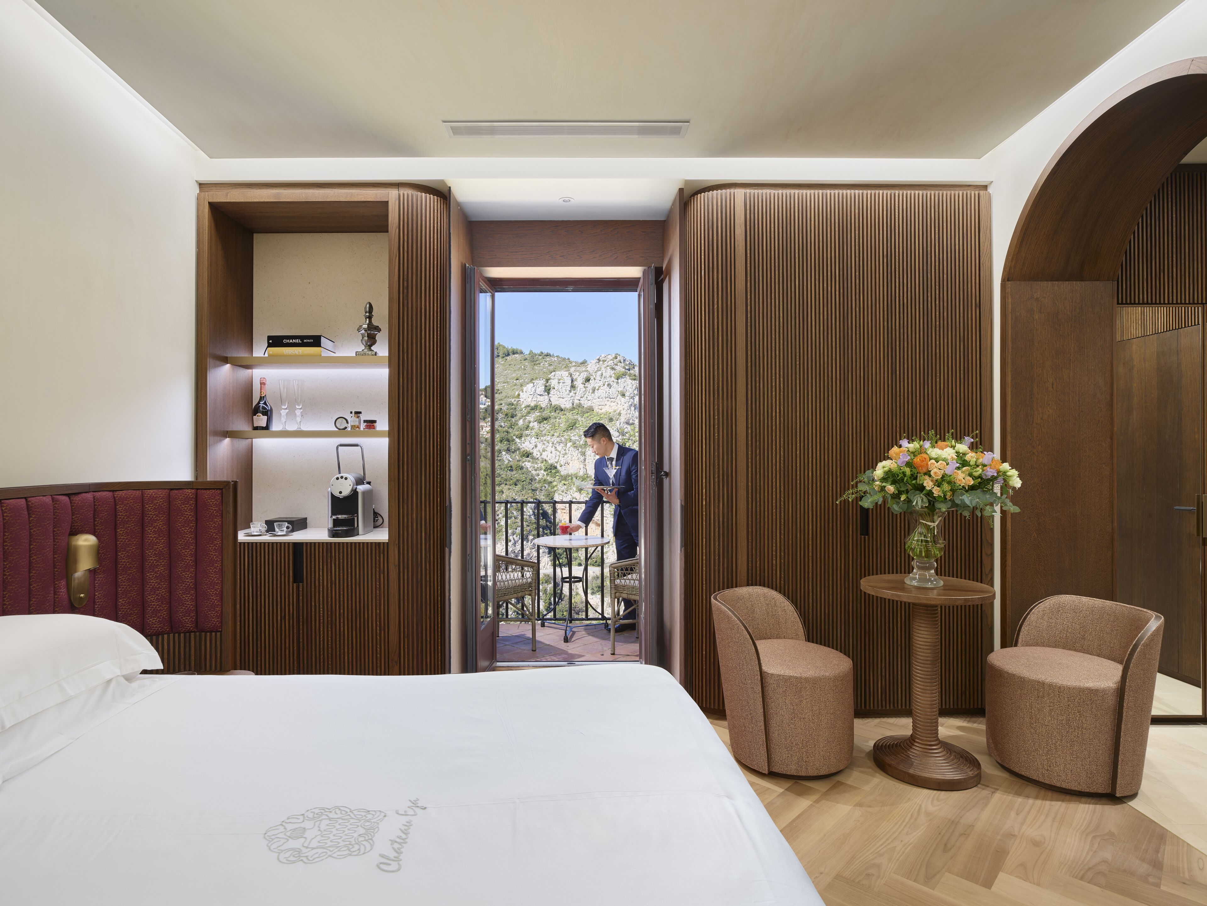 A hotel room with a bed, two armchairs, a small table, a vase with flowers, a minibar, and a balcony with a view of the mountains.