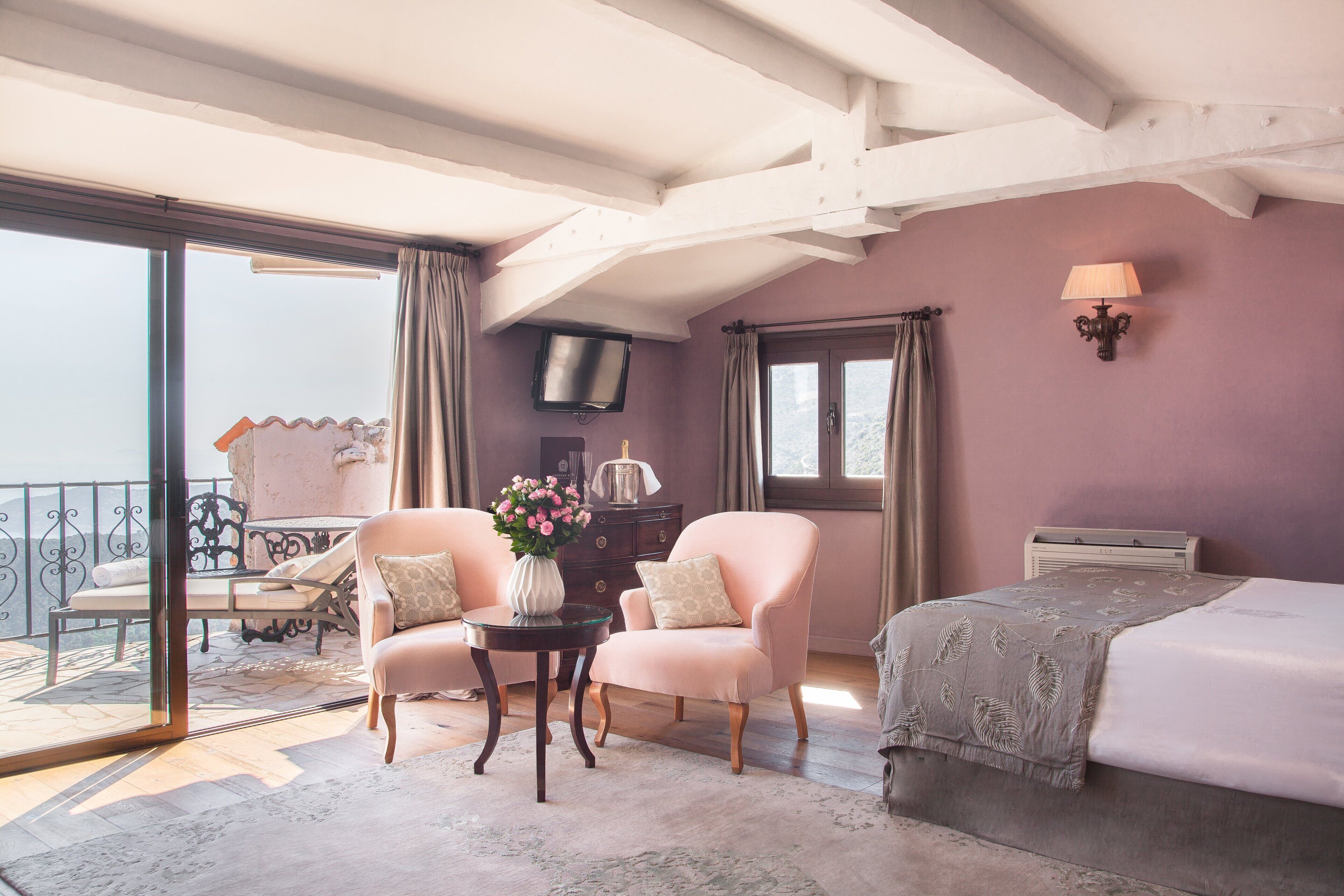 A room with a bed, two chairs, a small table with a vase of flowers, a television on the wall, and a balcony with a table and chairs.