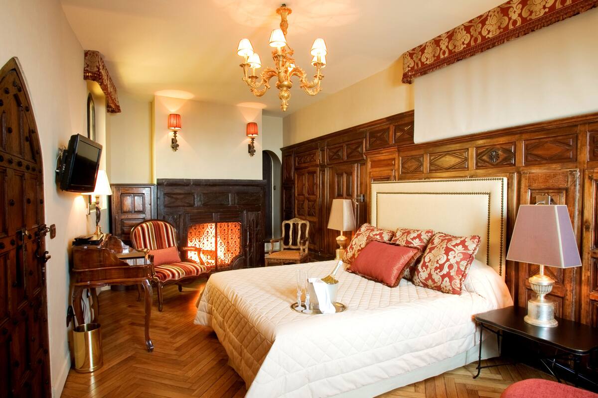 A hotel room with a large bed, a wooden headboard, a chandelier, a fireplace, and a television on a wooden cabinet.