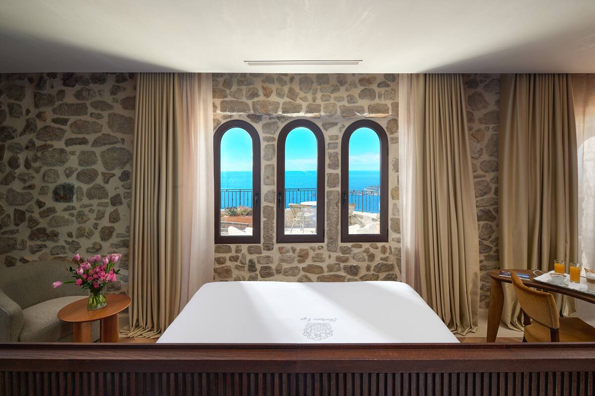 A hotel room with a stone wall, three arched windows with a view, a bed, a small table with a vase of flowers, and a dining area with a table and chairs.