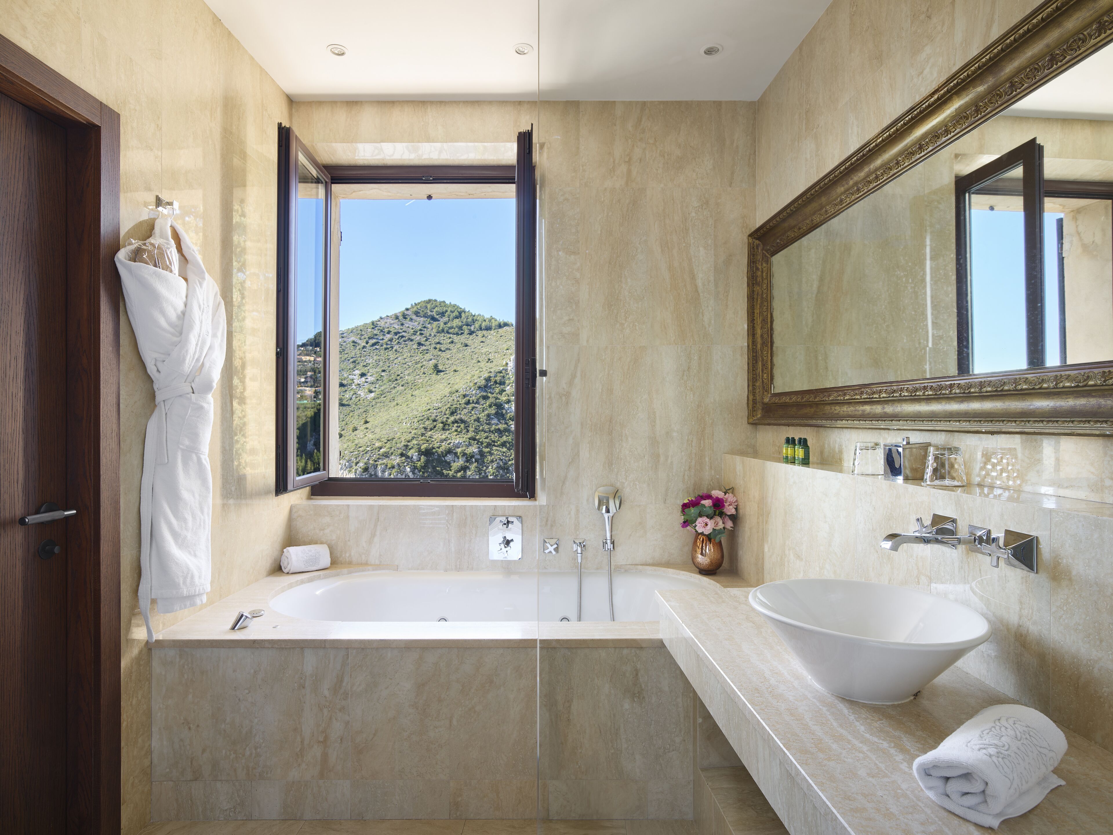 A modern bathroom with a large bathtub, a sink, and a window offering a view of a green hillside.