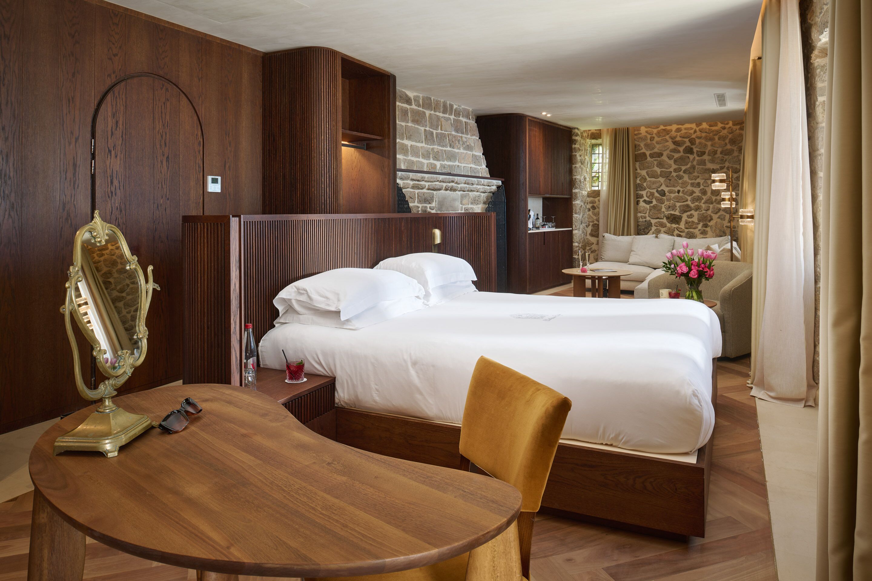 A hotel room with a large bed, a wooden table, a chair, a mirror, and a stone wall.
