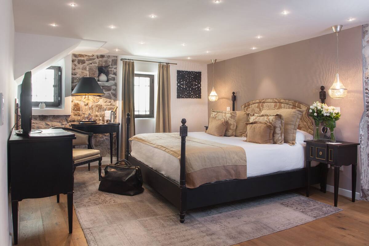 A bedroom with a stone wall, a bed with a headboard, a desk, a chair, and a nightstand with a lamp.